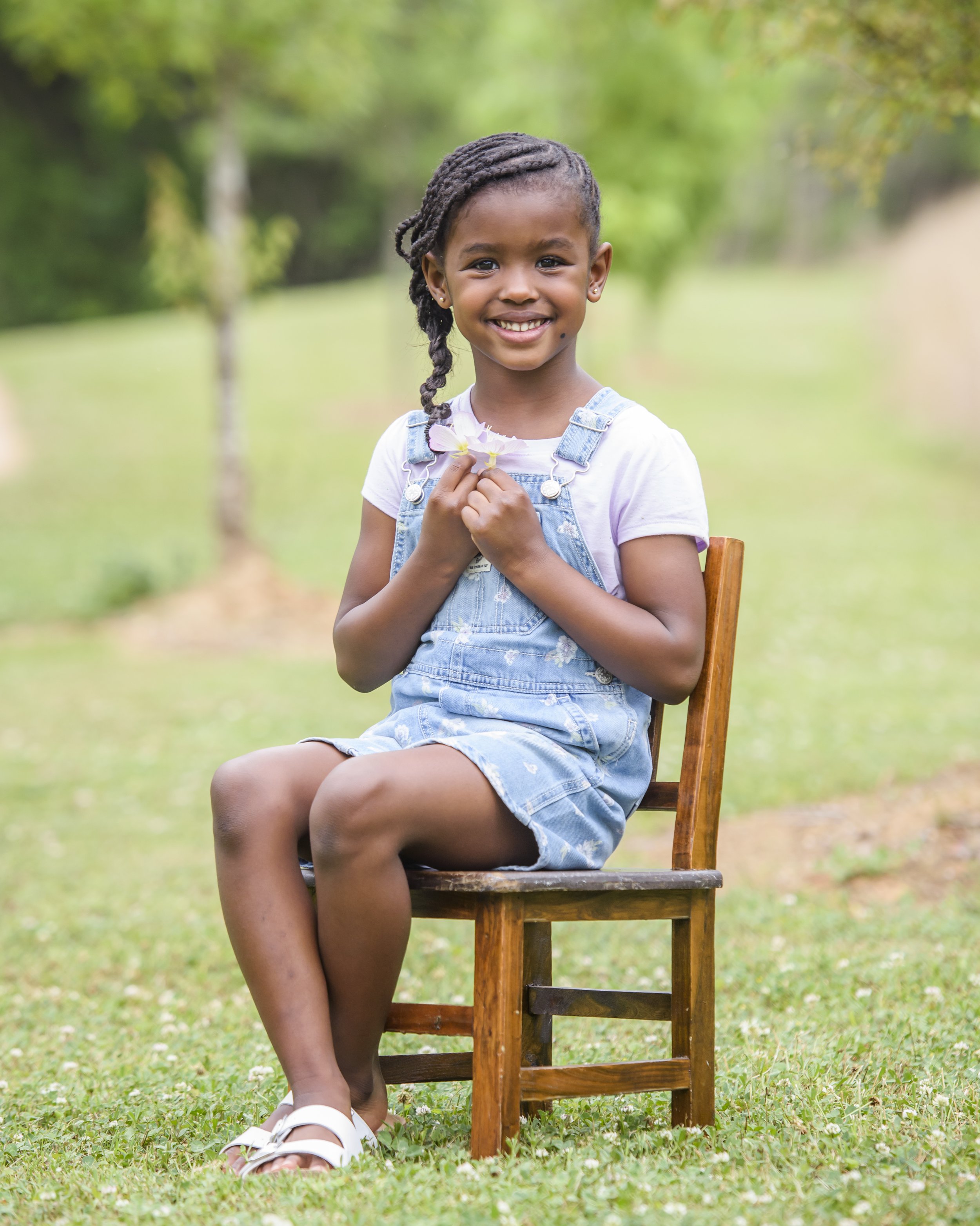 A young girl with braided hair, wearing a white t-shirt and denim overalls, sitting on a wooden chair outdoors in a grassy area with trees in the background. She is smiling and holding a small flower.