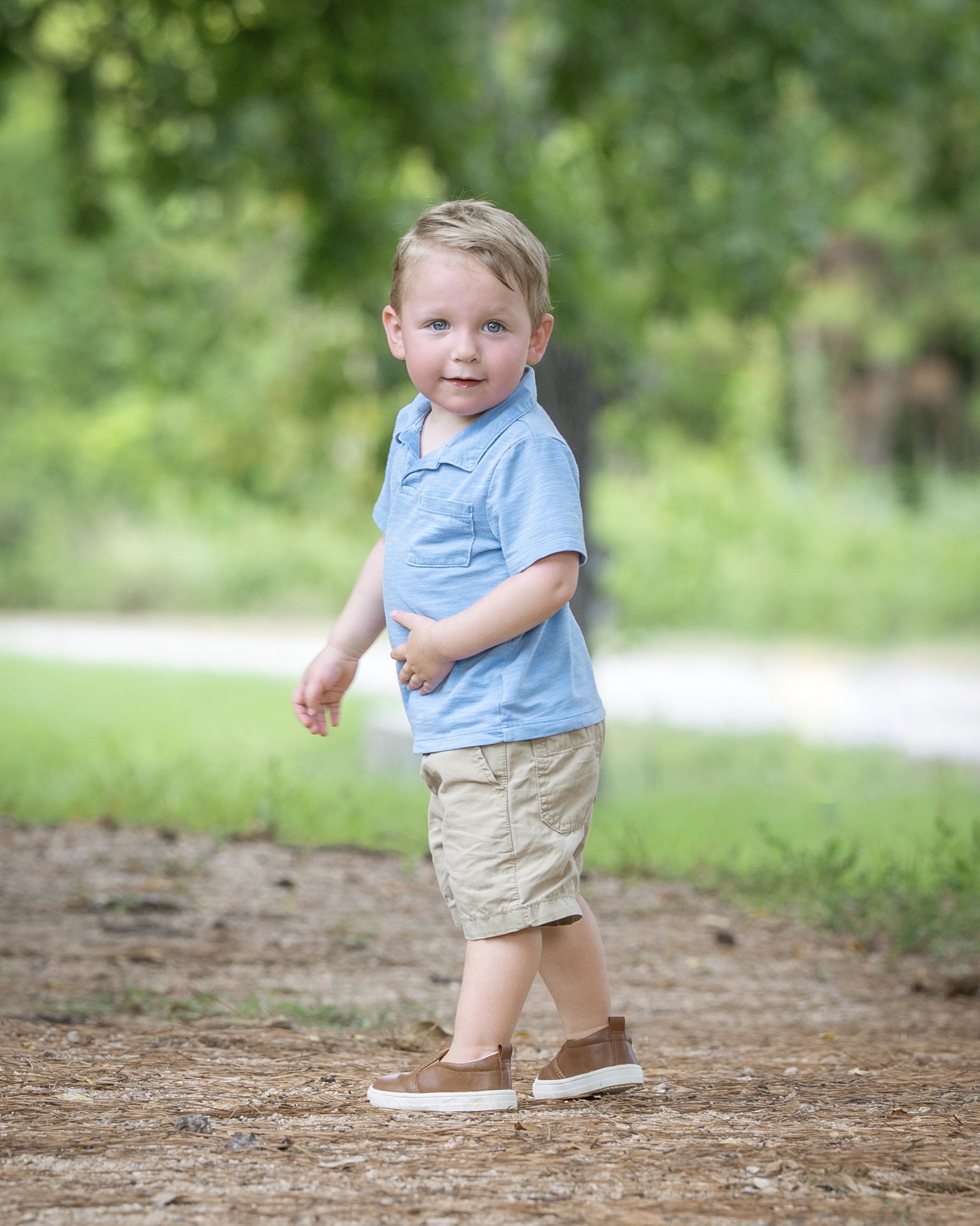 A young boy with blond hair and blue eyes standing outdoors on a dirt path, wearing a light blue polo shirt, tan shorts, and brown shoes with white soles, surrounded by green trees.