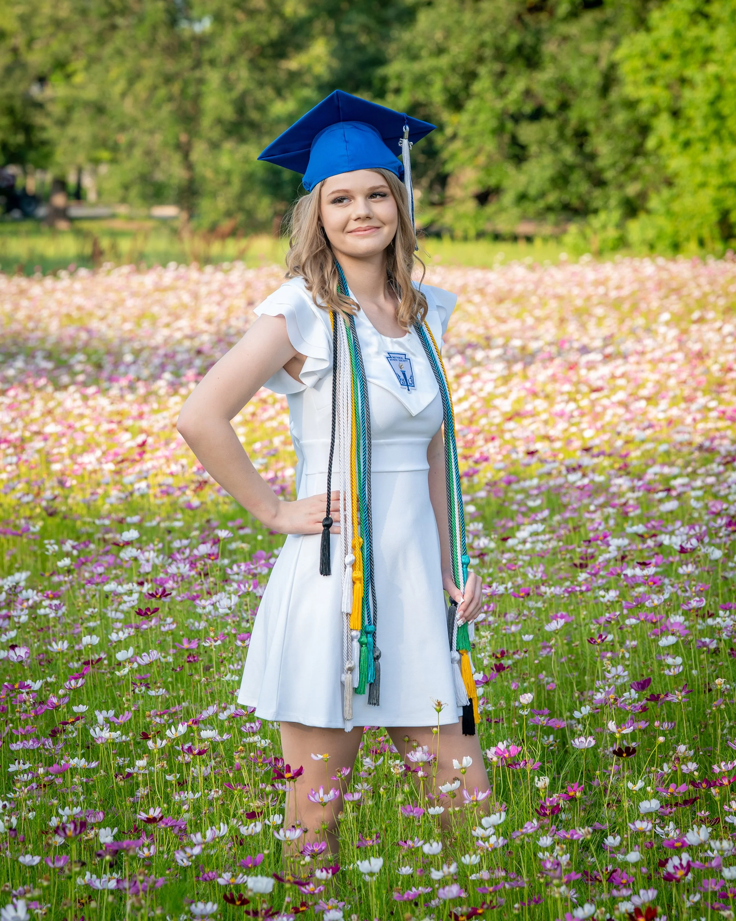 Young woman in a white dress and blue graduation cap and gown standing in a field of pink and white flowers with trees in the background.