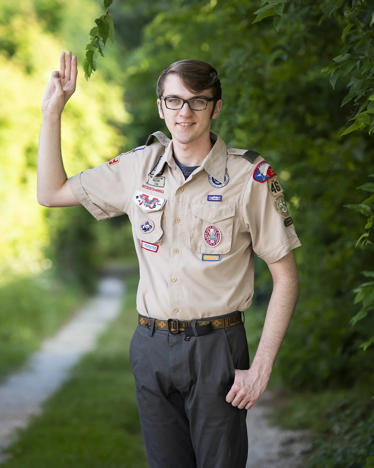 A young man in a Boy Scouts uniform standing on a nature trail, smiling and waving, surrounded by green trees.