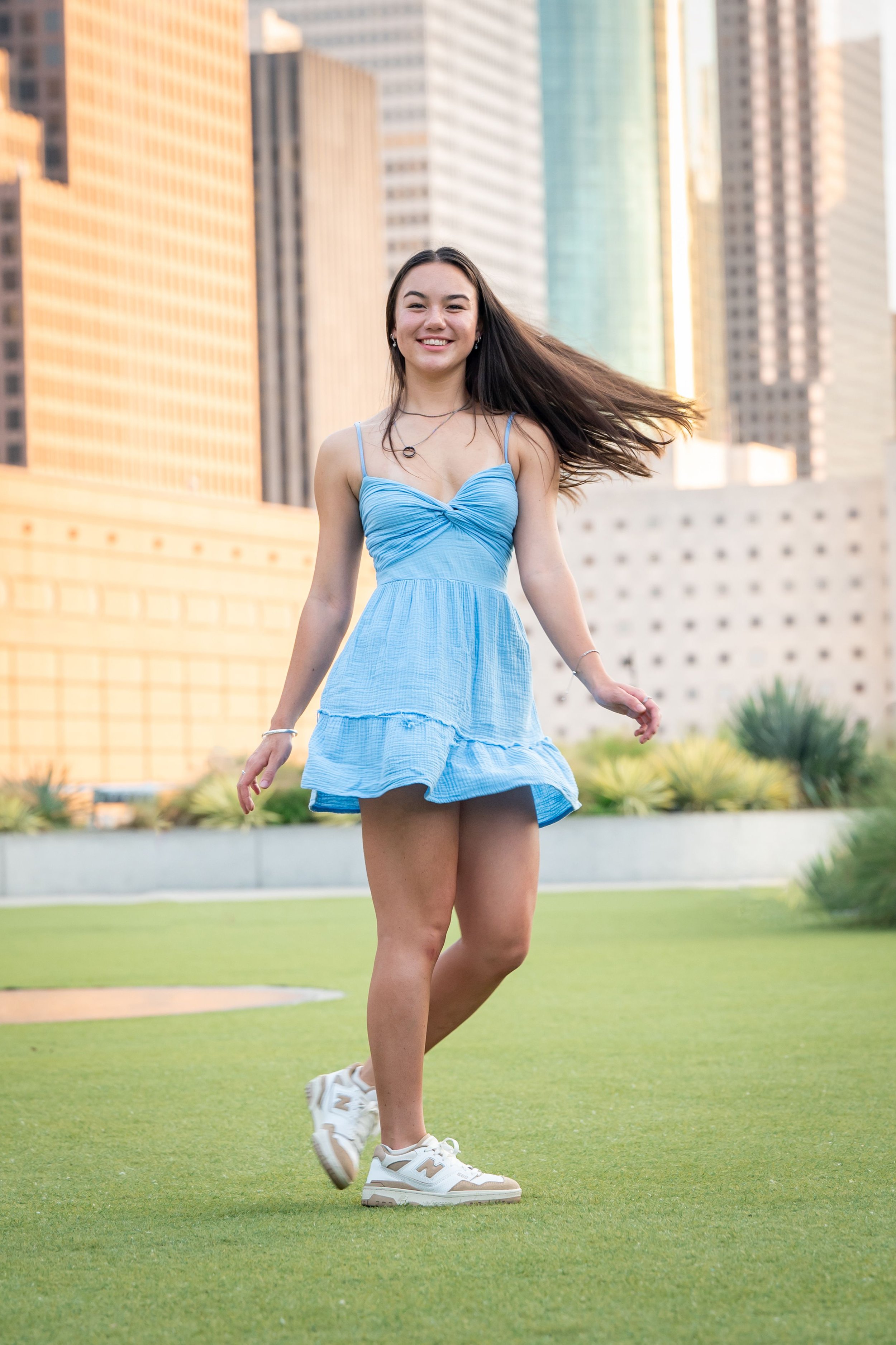 A senior girl in a blue dress walking on a grassy area in an urban park, surrounded by tall buildings.