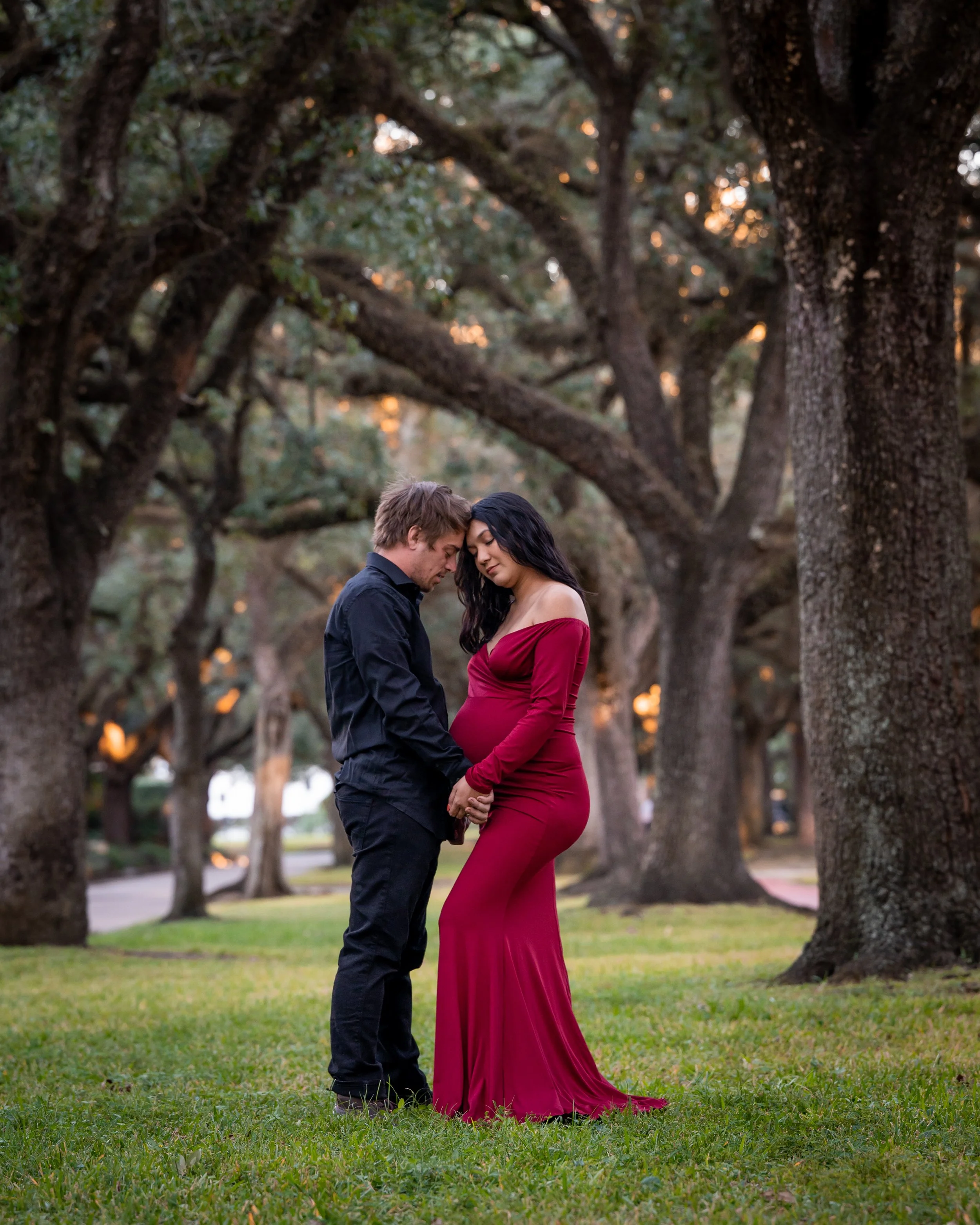 A couple standing close together in a park with large, sprawling trees in the background. The woman is wearing a long, off-the-shoulder, burgundy dress that accentuates her pregnant belly, and the man is dressed in a black shirt and pants. They are t