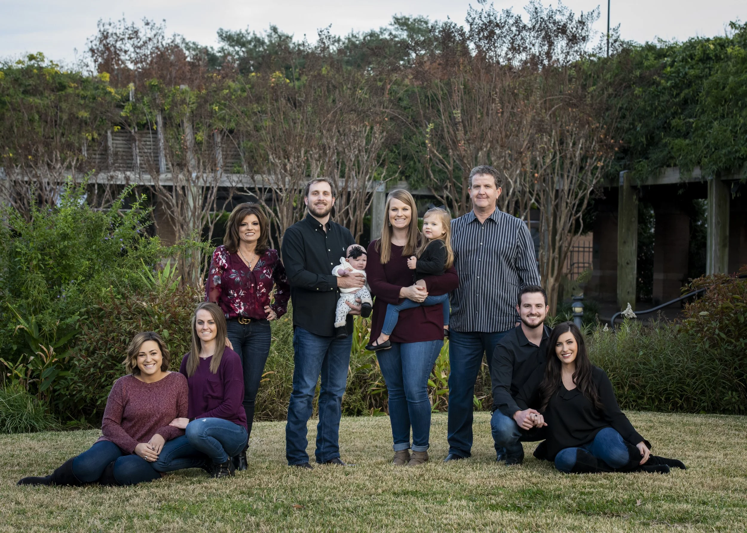 Family group photo outdoors in a garden with trees and bushes, featuring women, men, and children smiling and posing for the camera.