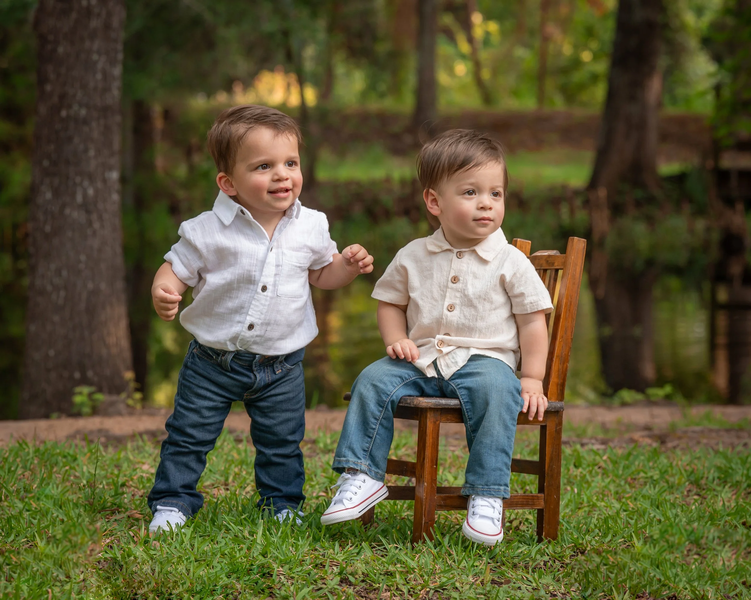 Two young boys with brown hair, one standing and one sitting on a wooden chair, in a wooded outdoor setting with trees and greenery, during daytime.