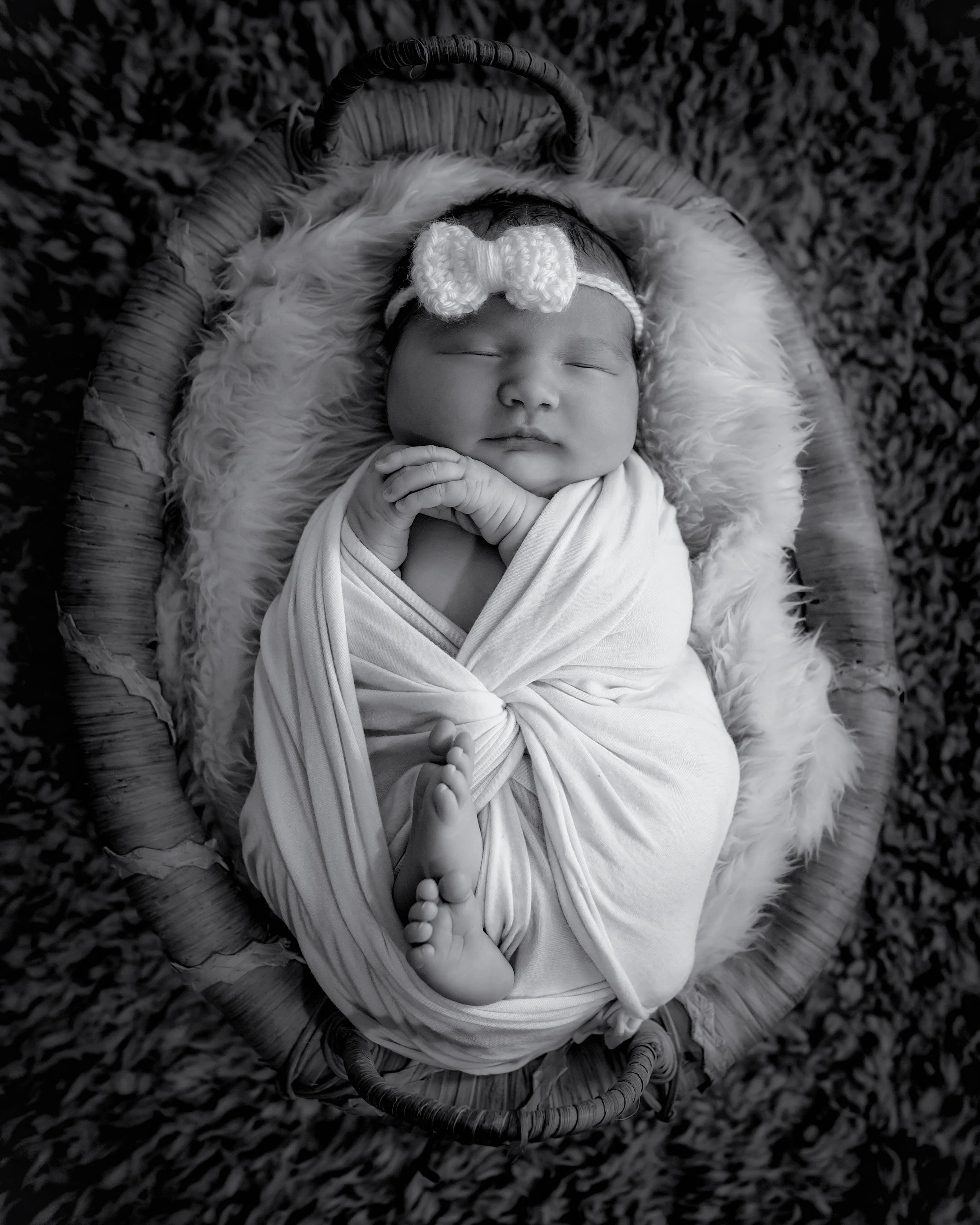 A newborn baby in a woven basket on a soft fur blanket, wearing a knitted bow headband and wrapped in a cloth, with one hand near the face and the other resting on the chest, surrounded by a textured background.