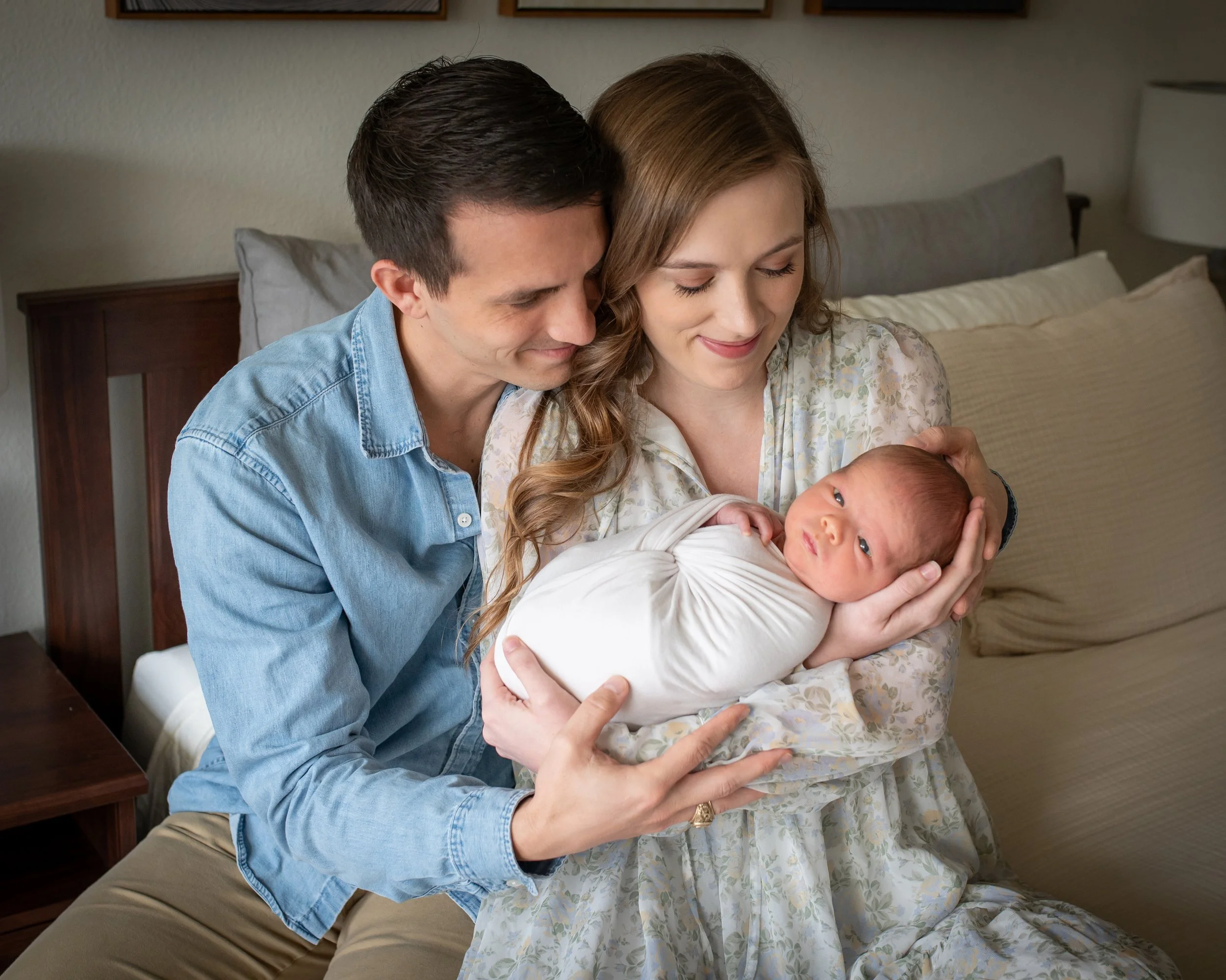 A man and a woman are sitting on a bed, holding and looking at a newborn baby. They appear happy and are gazing at the baby lovingly. The woman has long, curly hair and is wearing a floral dress, while the man has short, dark hair and is wearing a de