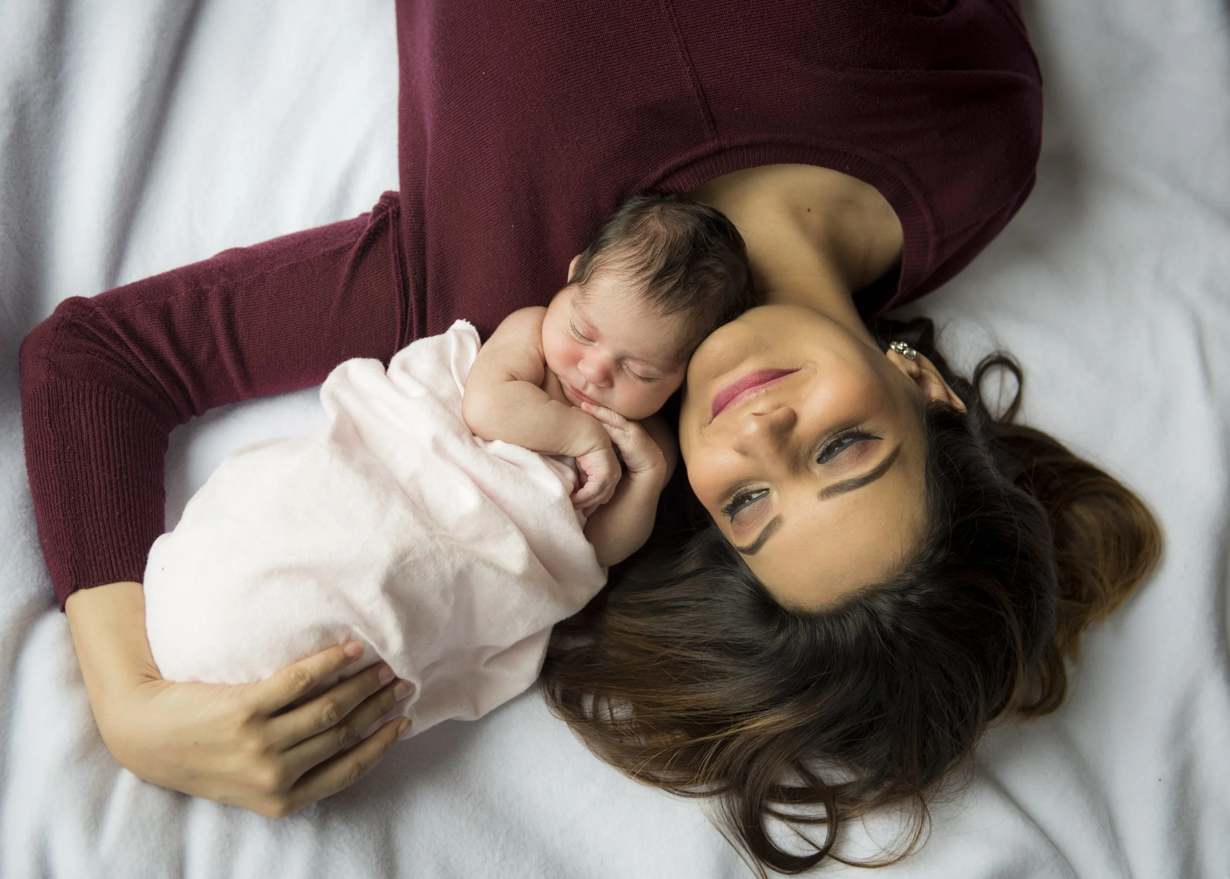 A woman with long brown hair lying on her back with a newborn baby resting on her chest, both smiling and appearing happy.