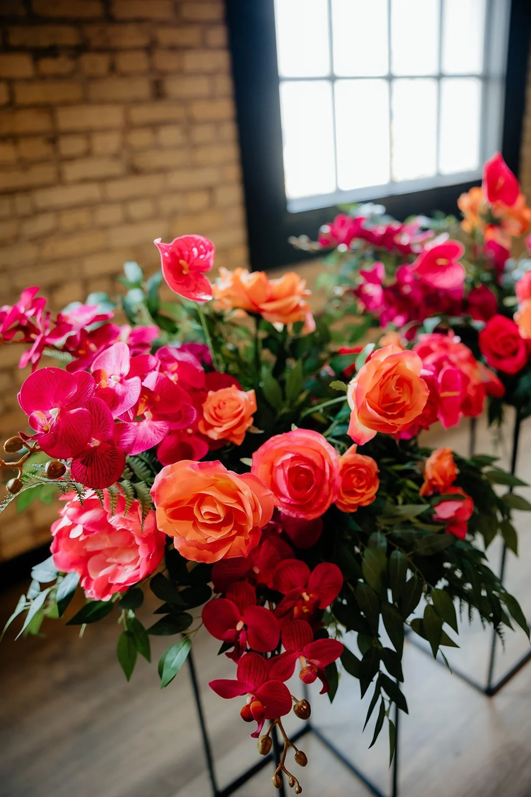 A bouquet of colorful flowers, including roses and orchids, arranged in front of a brick wall and a window.