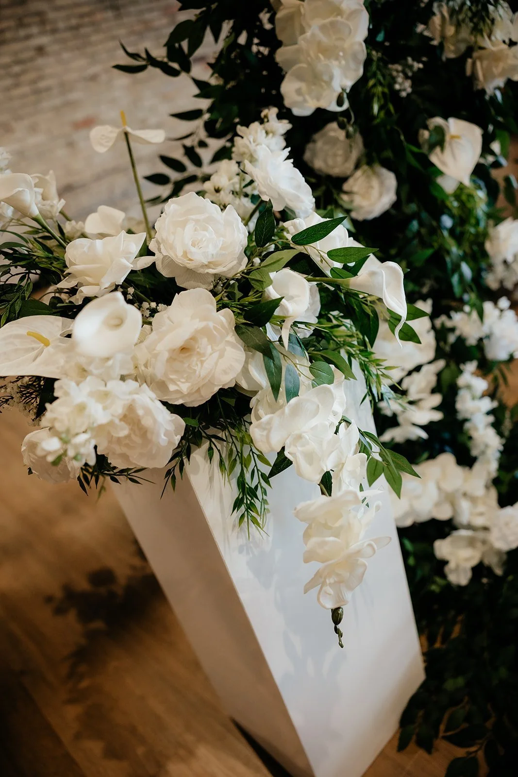 White floral arrangement with roses and other flowers in a white square vase, on a wooden surface.