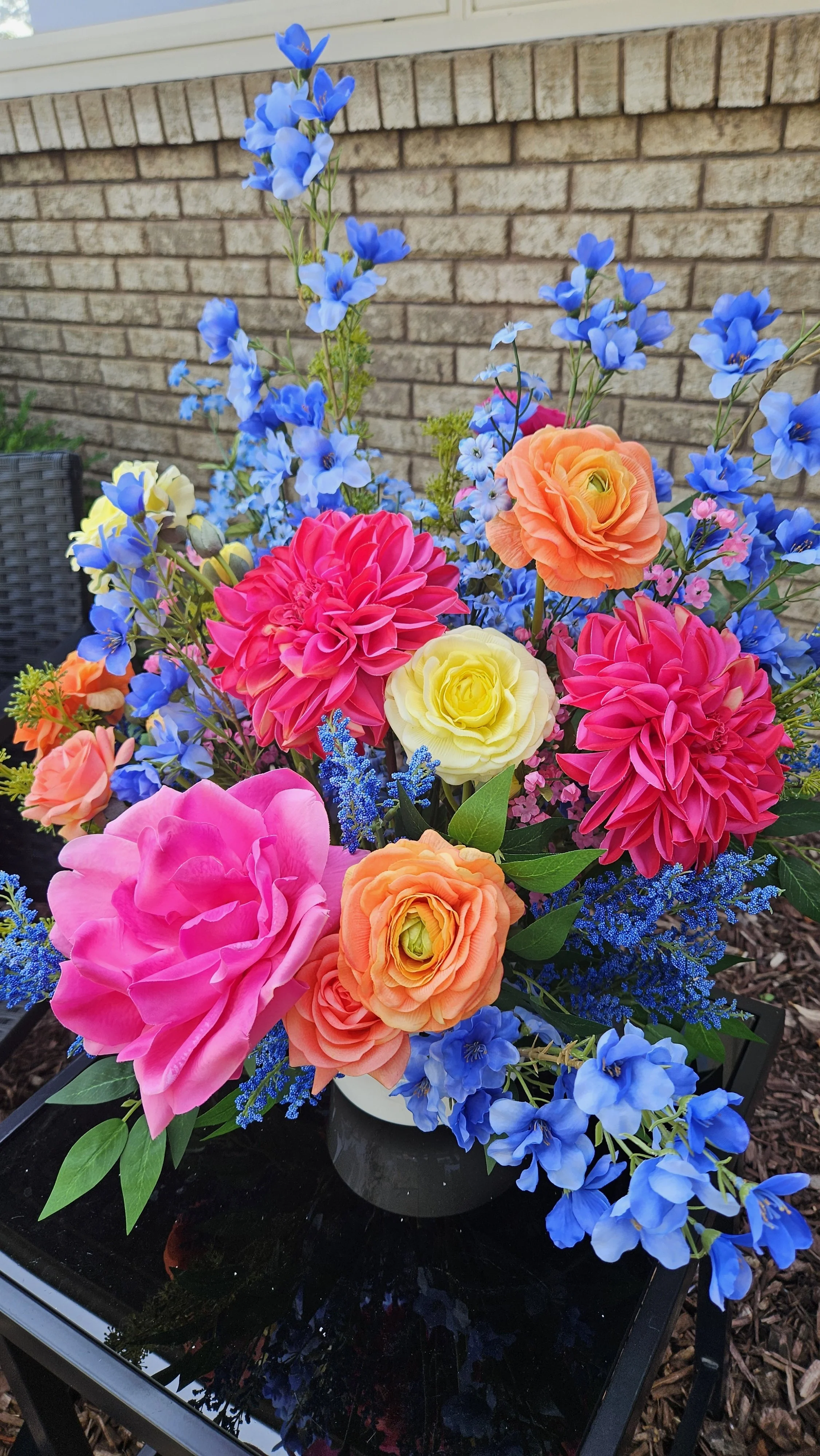 Colorful flower arrangement featuring pink dahlias, yellow roses, orange ranunculus, and blue delphiniums on a black table against a brick wall.