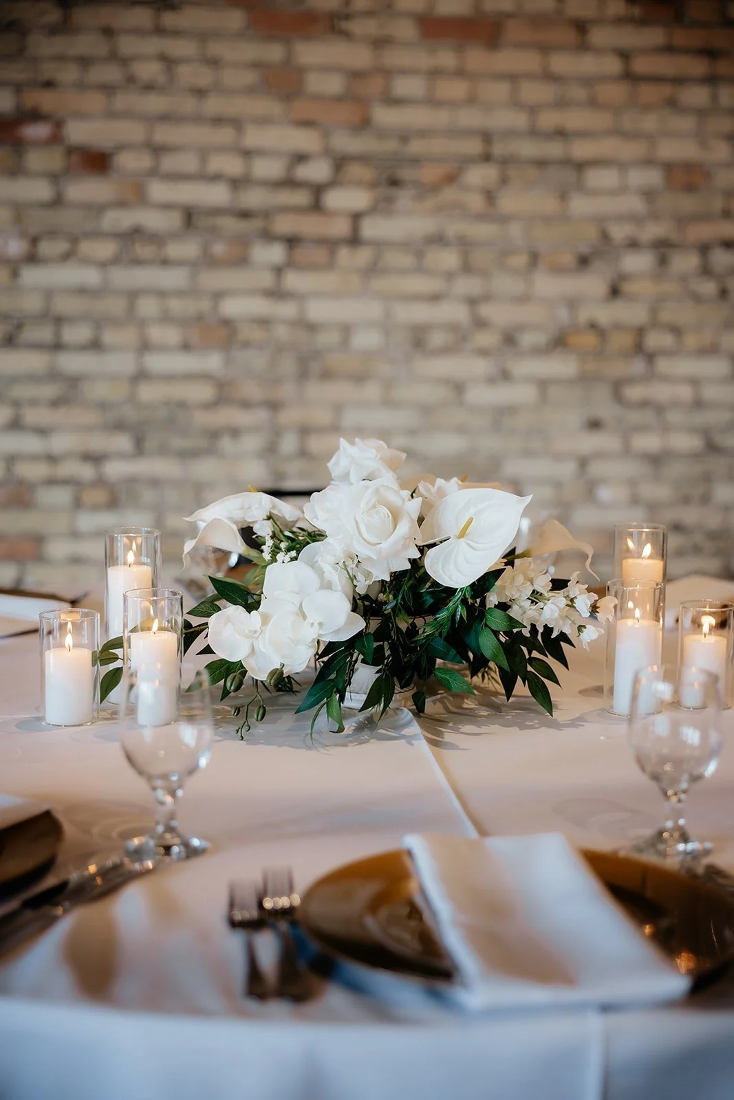 Elegant table setting with a white floral centerpiece, white candles in glass holders, and glassware, against a brick wall background.
