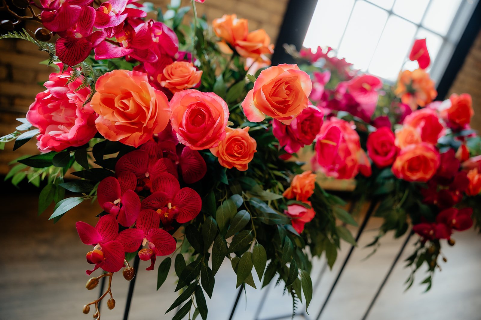 Arrangement of pink, orange, and red roses with red orchids and green leaves.