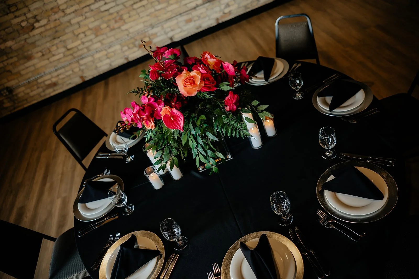 A round table set for a formal dinner with black tablecloths, black napkins, white plates with gold-rimmed chargers, wine glasses, forks, knives, and spoons. In the center, there is a large floral arrangement with pink and peach roses, pink anthurium