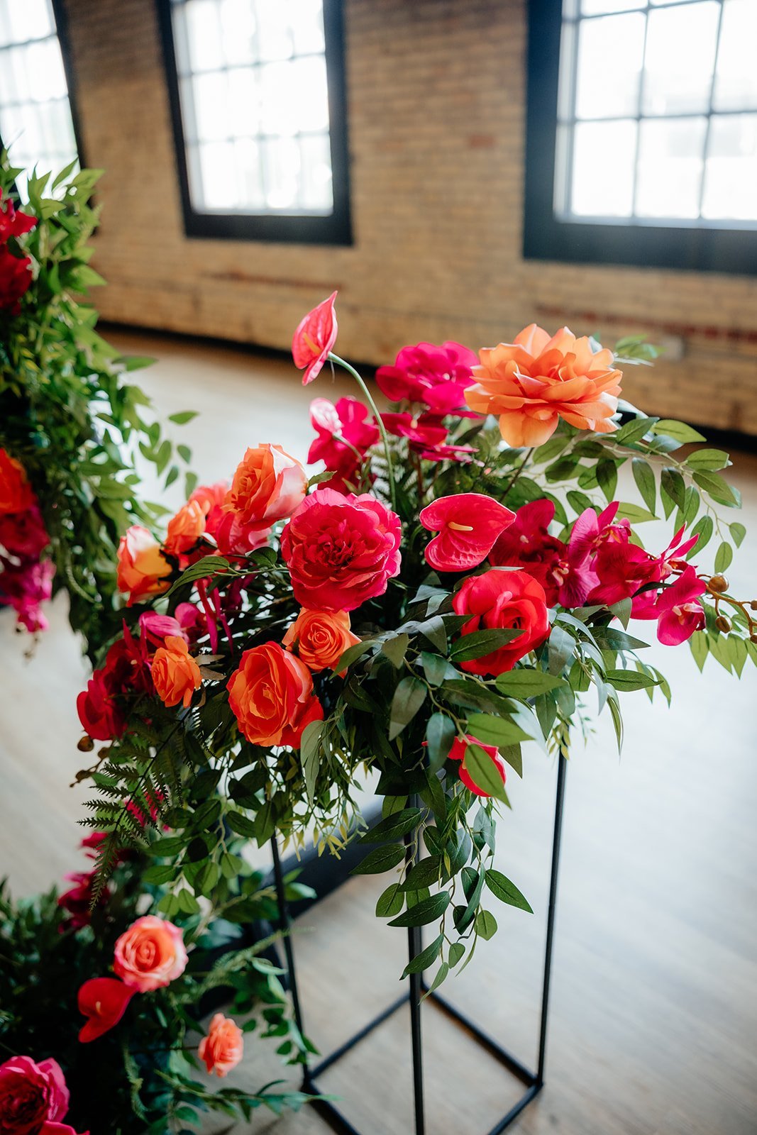 Colorful arrangement of pink, orange, and red flowers with green leaves in an indoor setting with large windows and brick walls.
