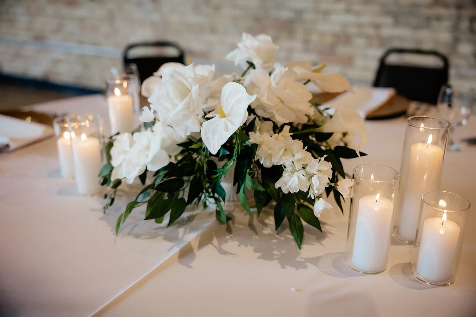 Elegant table centerpiece with white flowers and green leaves, surrounded by lit white candles in glass holders, set on a white tablecloth in a decorated indoor setting.