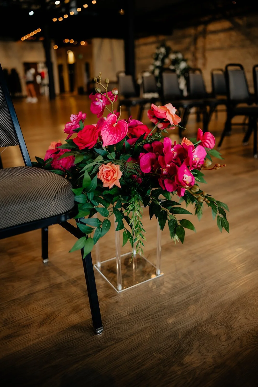 A large bouquet of pink and orange flowers, including roses and orchids, in a clear rectangular vase on a wooden floor. Chairs are visible in the blurred background of an indoor event space.