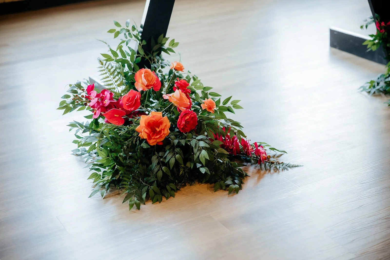 A floral arrangement with orange, red, and pink flowers along with green leaves and ferns on a wooden floor.