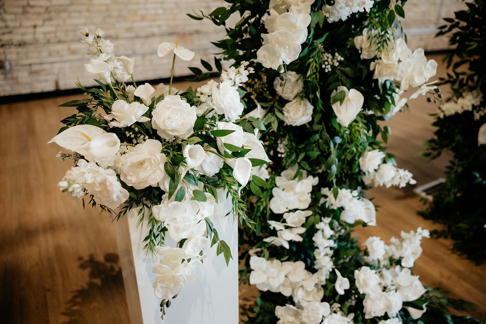 White floral arrangement with roses and calla lilies, with green leaves, set against a brick and wood background.