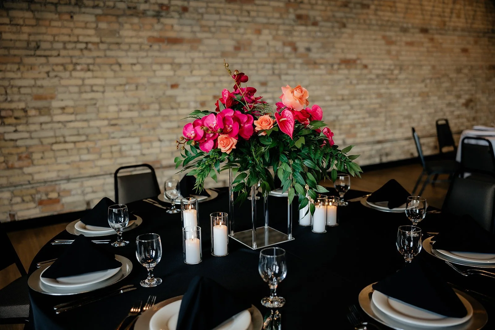 A dining table set for a formal event with a large floral centerpiece consisting of pink and peach roses, pink orchids, and green foliage. Surrounding the arrangement are black napkins, white plates, silverware, empty wine glasses, and white candles 