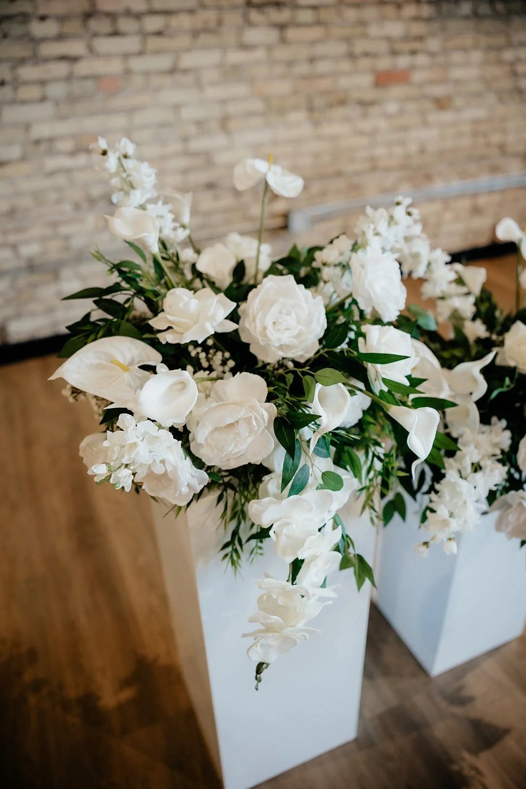 A white vase filled with a lush arrangement of white flowers, including roses, calla lilies, and other blooms, accented with green leaves, placed on a wooden floor against an exposed brick wall.