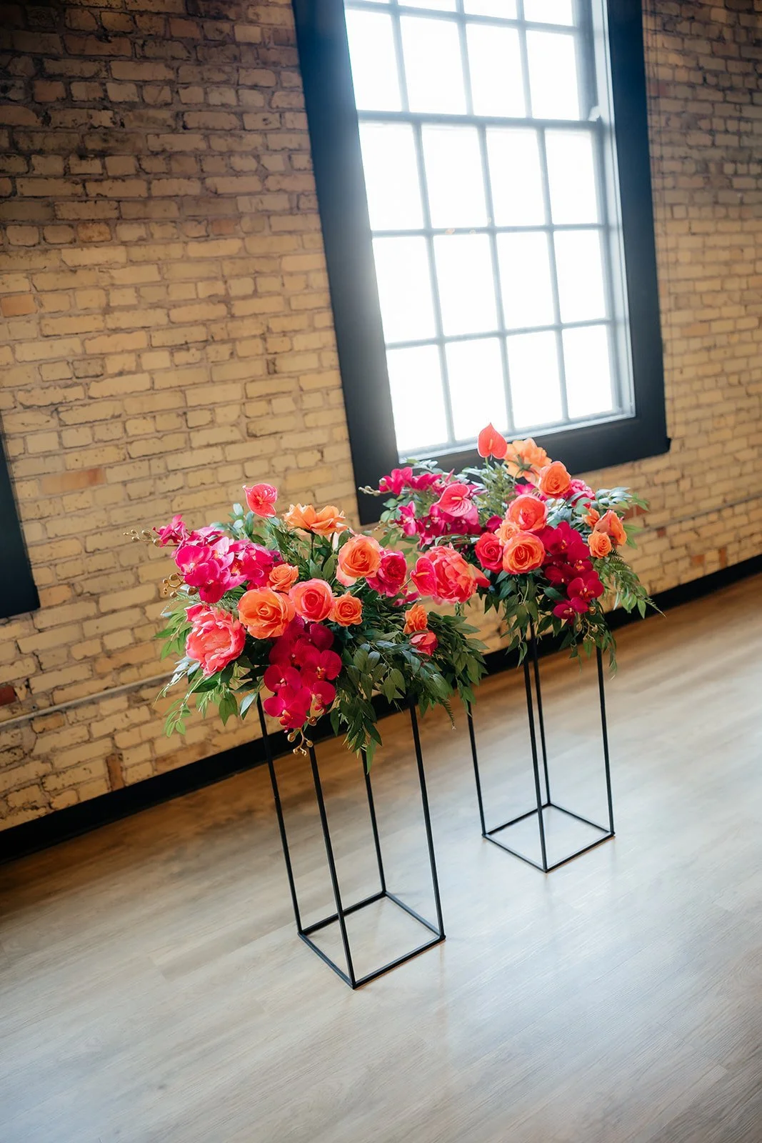 Two black metal plant stands holding pink and orange floral arrangements with greenery, placed in front of a large window with black trim and a brick wall background.