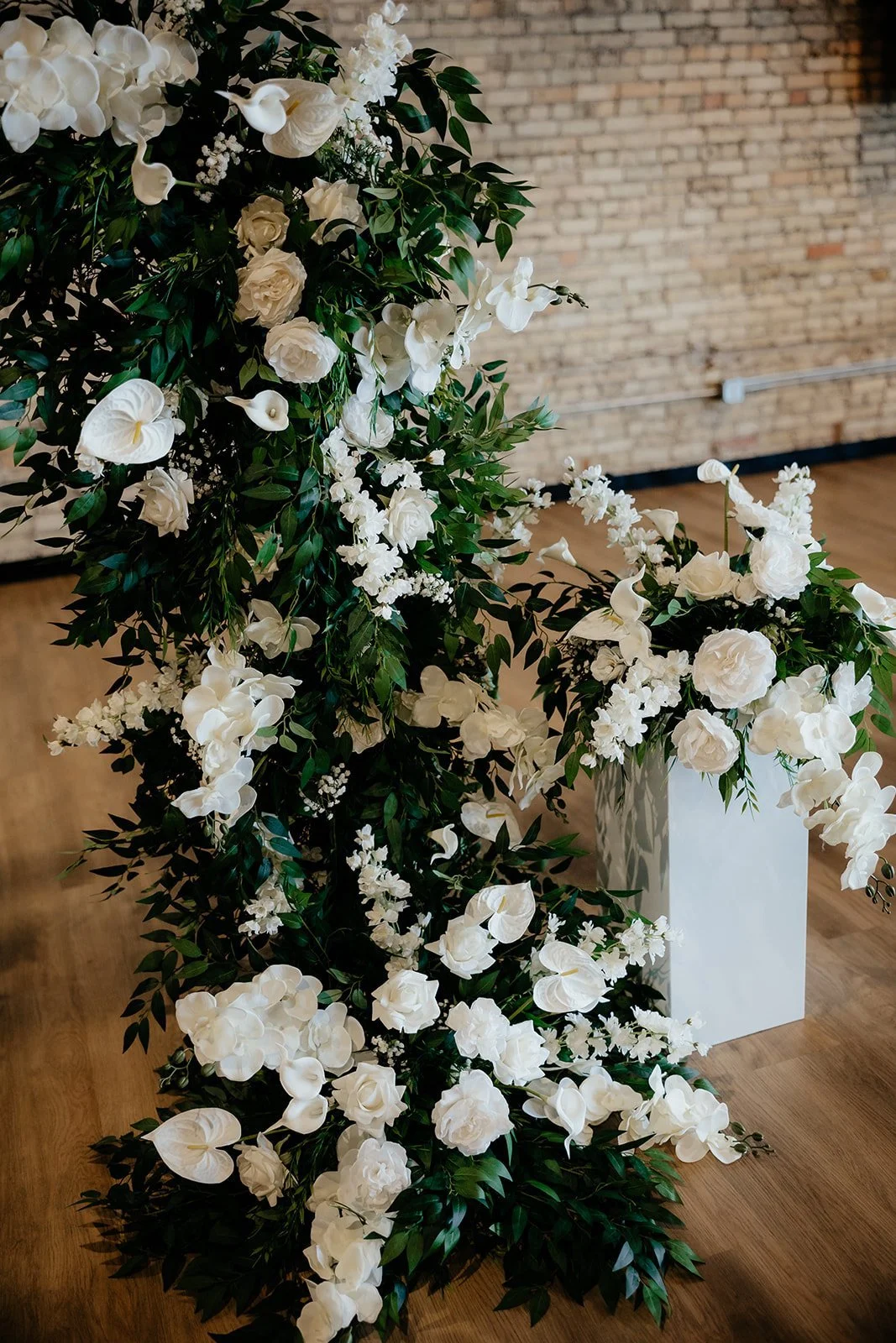 A large floral arrangement with white roses, calla lilies, and other white flowers complemented by green foliage, displayed on a white pedestal in front of a brick wall.
