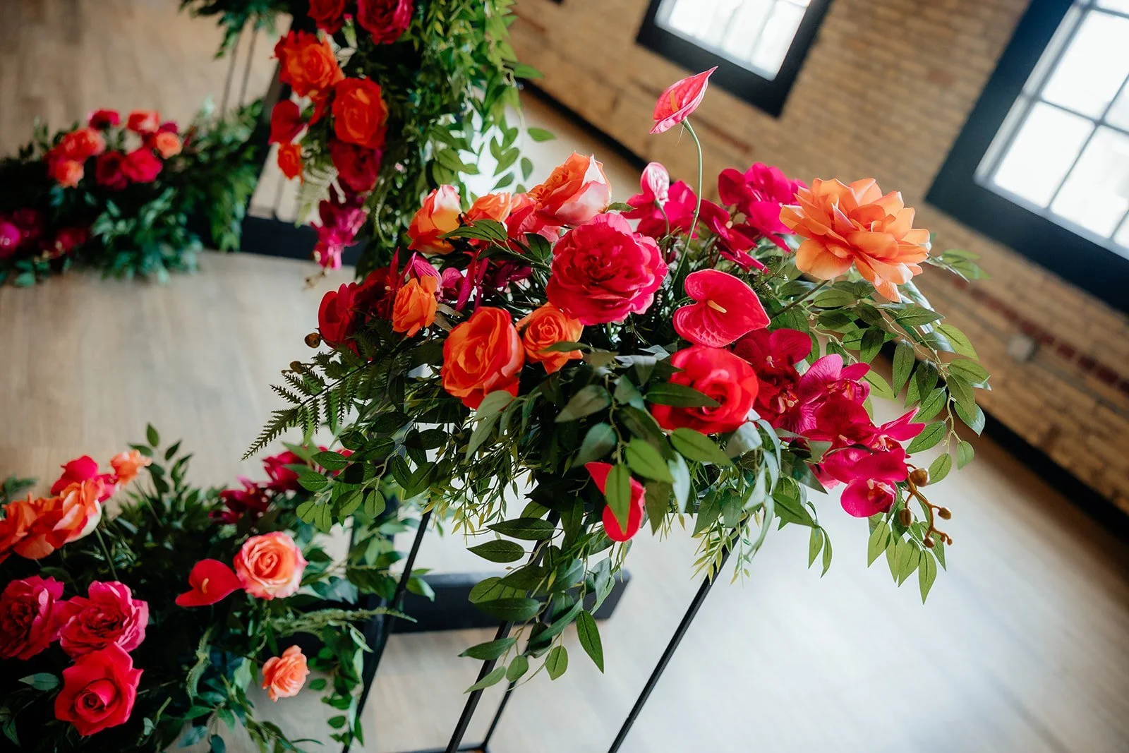 A vibrant bouquet of various pink, orange, and red flowers with green foliage, displayed on black metal stands inside a room with brick walls and large windows.