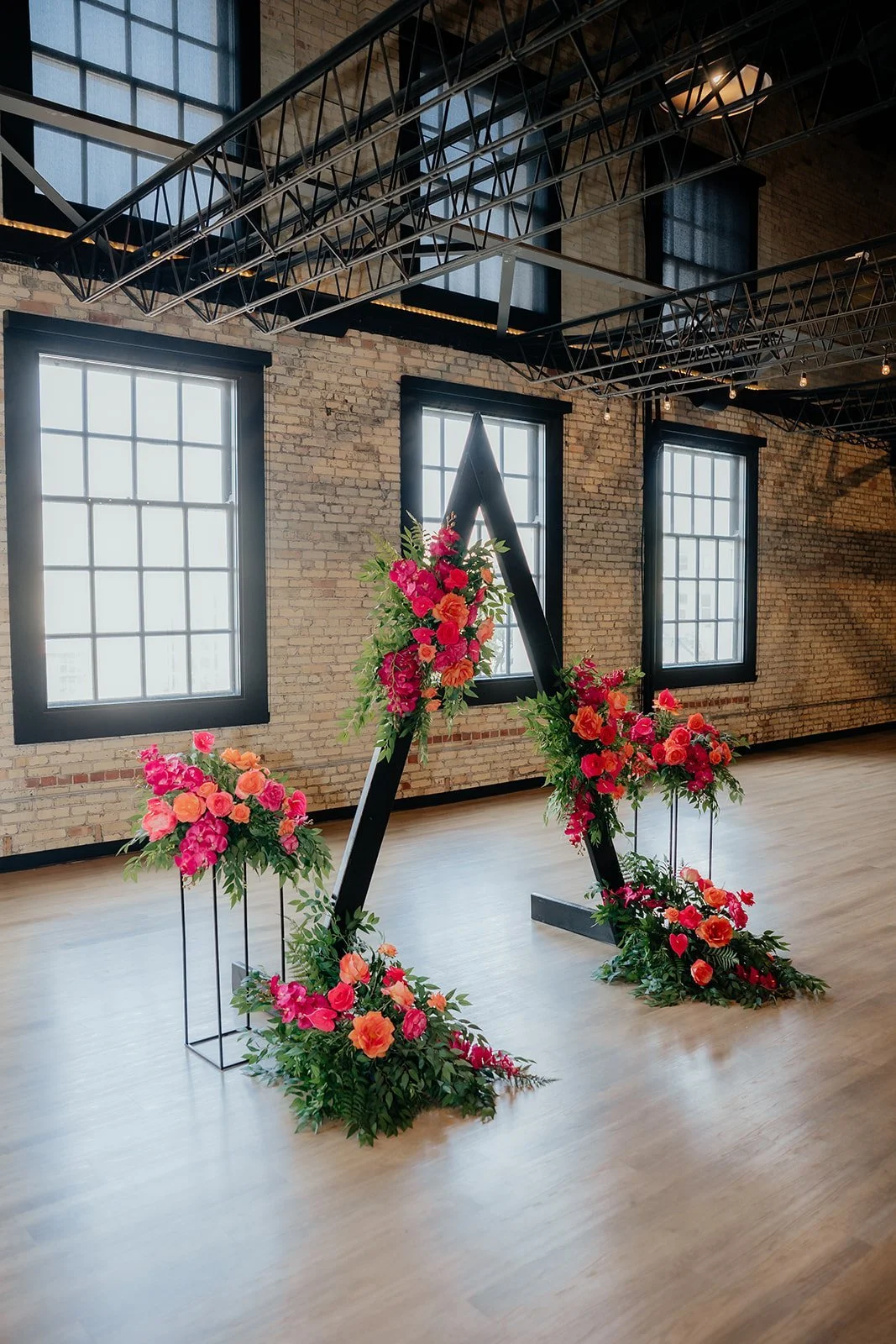 A floral arrangement in the shape of the letter A with pink and orange flowers and greenery, in a spacious industrial-style room with brick walls, large windows, and exposed ceiling beams.