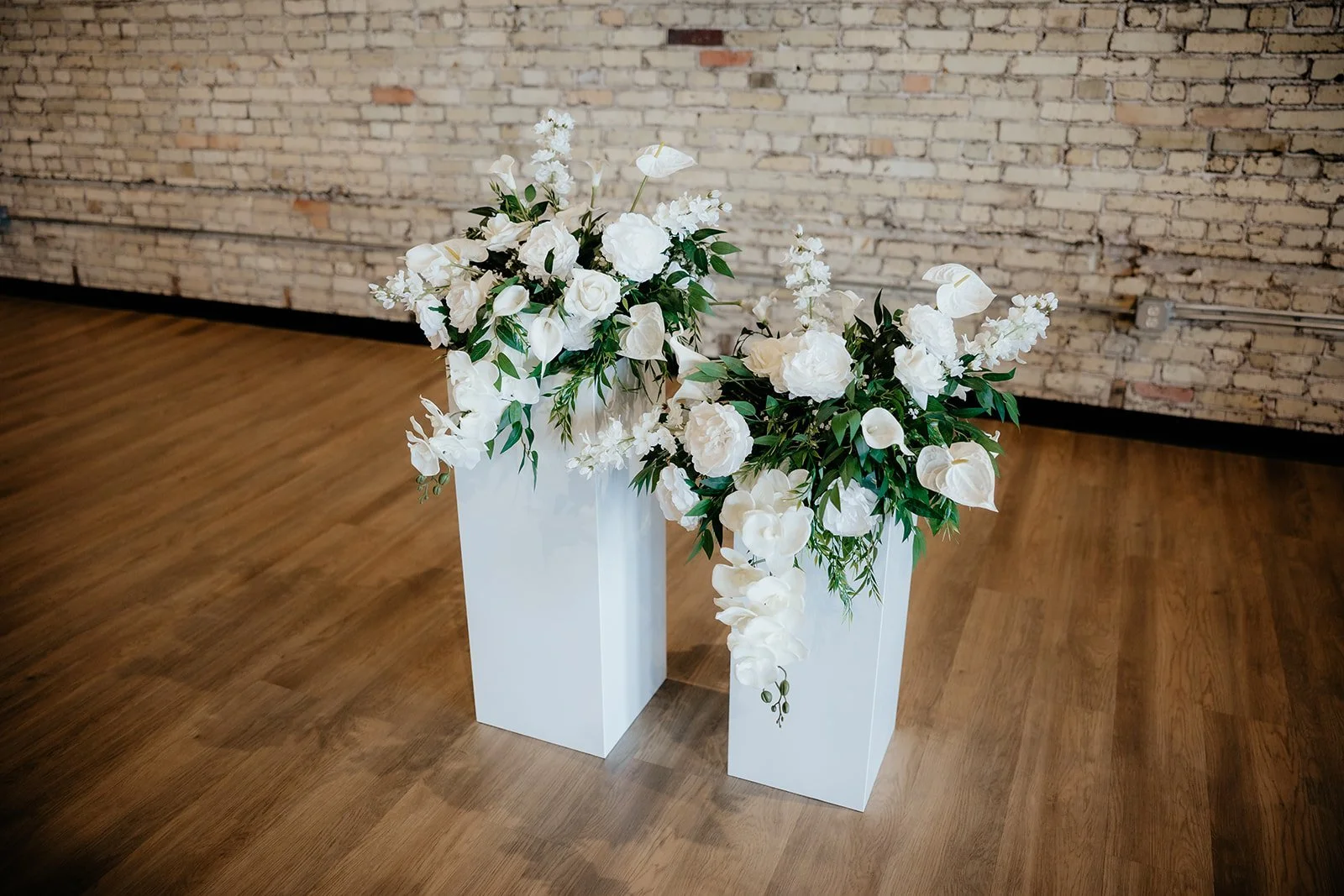 Two white rectangular pedestals with floral arrangements of white flowers and green leaves in a room with wood flooring and an exposed brick wall
