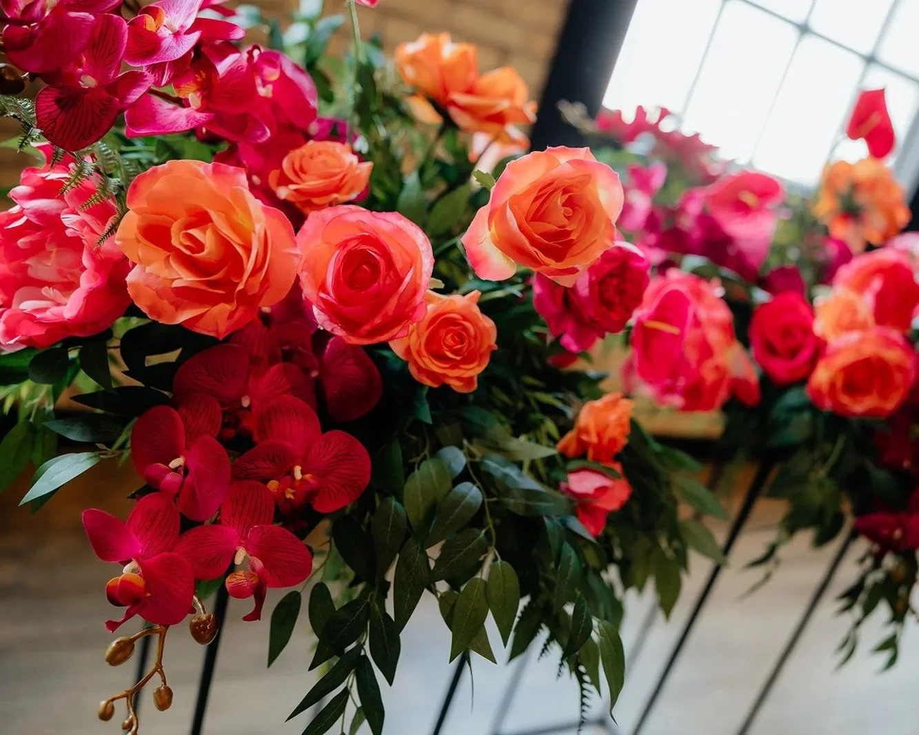 Close-up of vibrant orange, red, and pink roses and orchids in a floral arrangement, with a window in the background.
