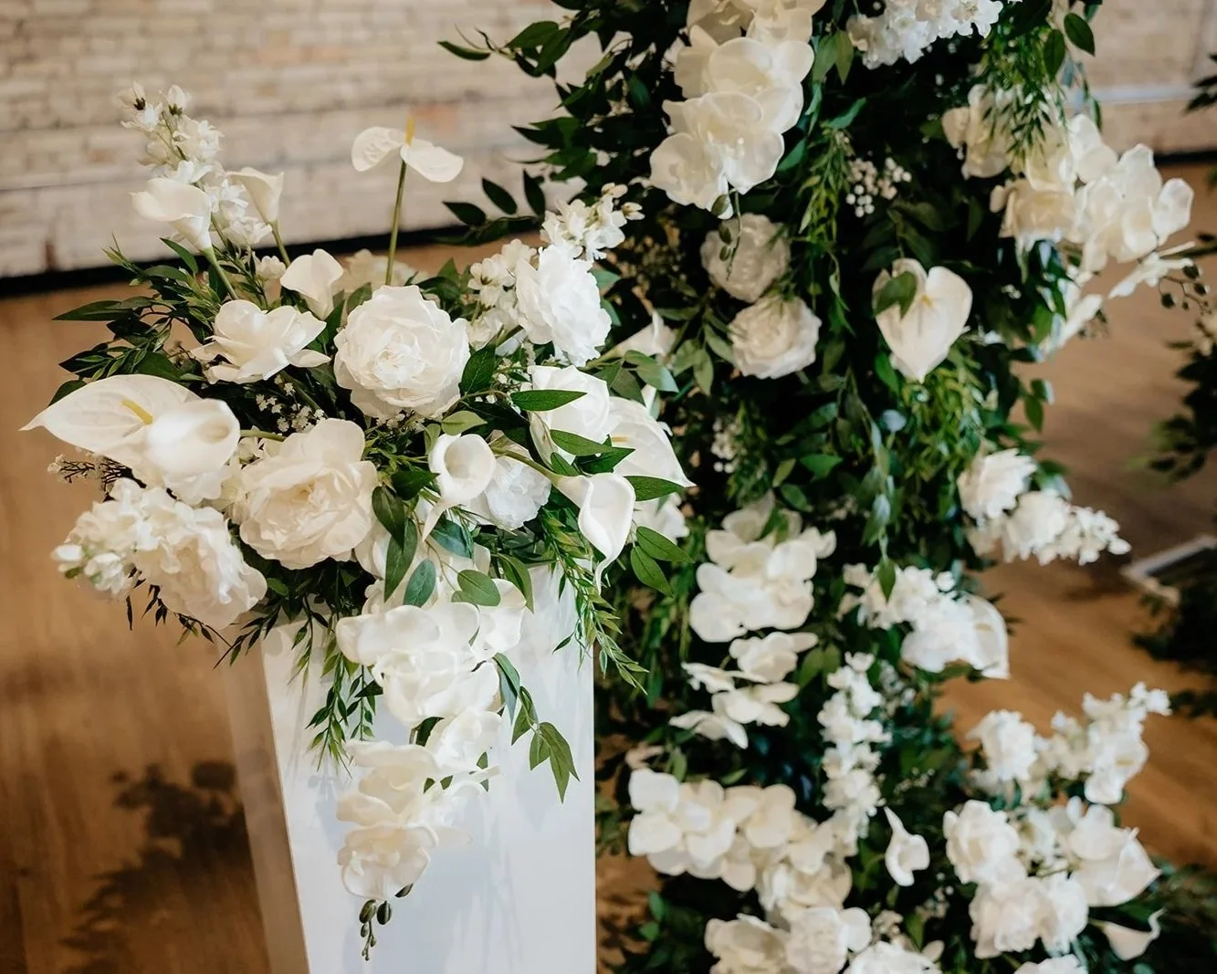 Close-up of a white floral arrangement with roses, calla lilies, and greenery, set against a wooden floor and brick wall.