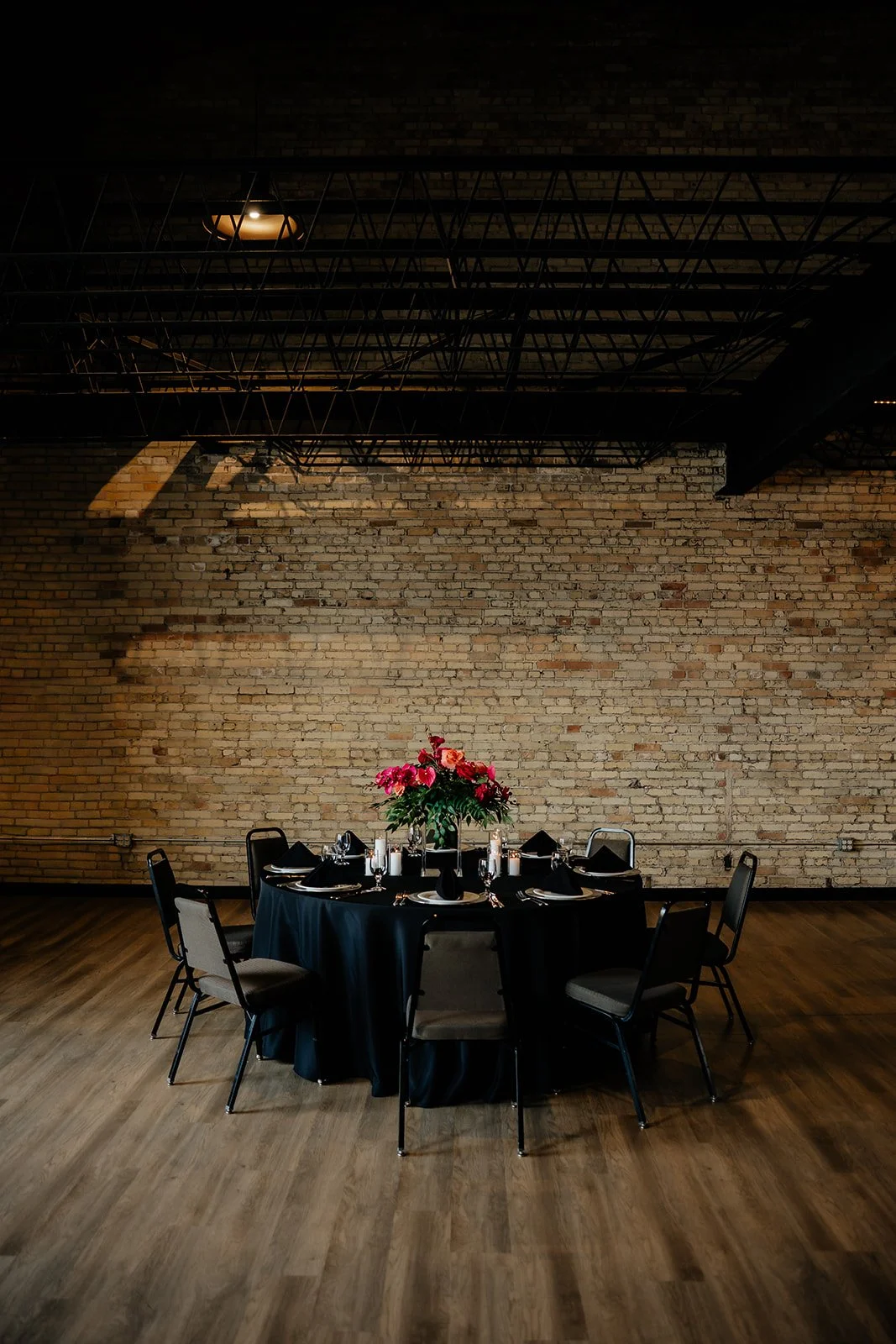 Round dining table with a black tablecloth, set with black napkins, candles, and a large pink floral centerpiece, surrounded by six chairs against an exposed brick wall in an industrial-style room.