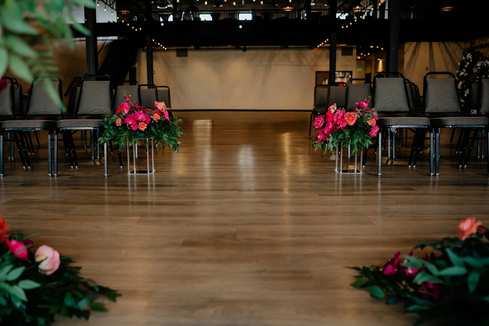 A wedding or event setup with black chairs lined up on the sides of a wooden floor, decorated with vibrant pink and orange flowers in silver stands, in a spacious indoor venue with warm lighting and string lights overhead.