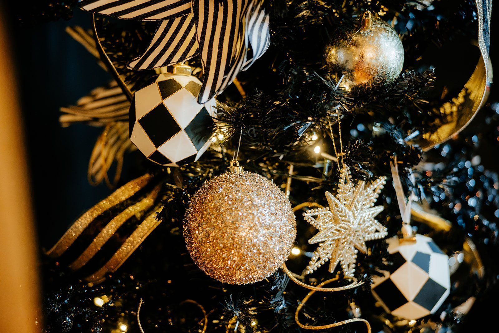 Close-up of a decorated Christmas tree with black, gold, and white ornaments, including a glittering gold ball, a geometric black and white ornament, a golden snowflake, and a shiny silver ball, illuminated by warm lights.