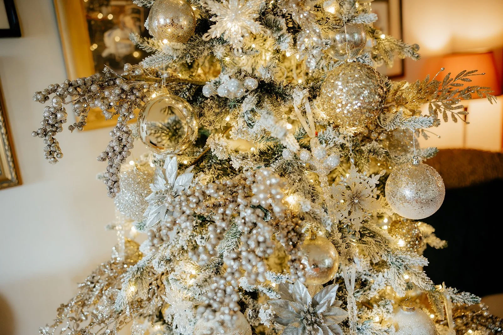 Close-up of a decorated Christmas tree with gold and silver ornaments, including baubles, snowflakes, and beads, with warm white lights.