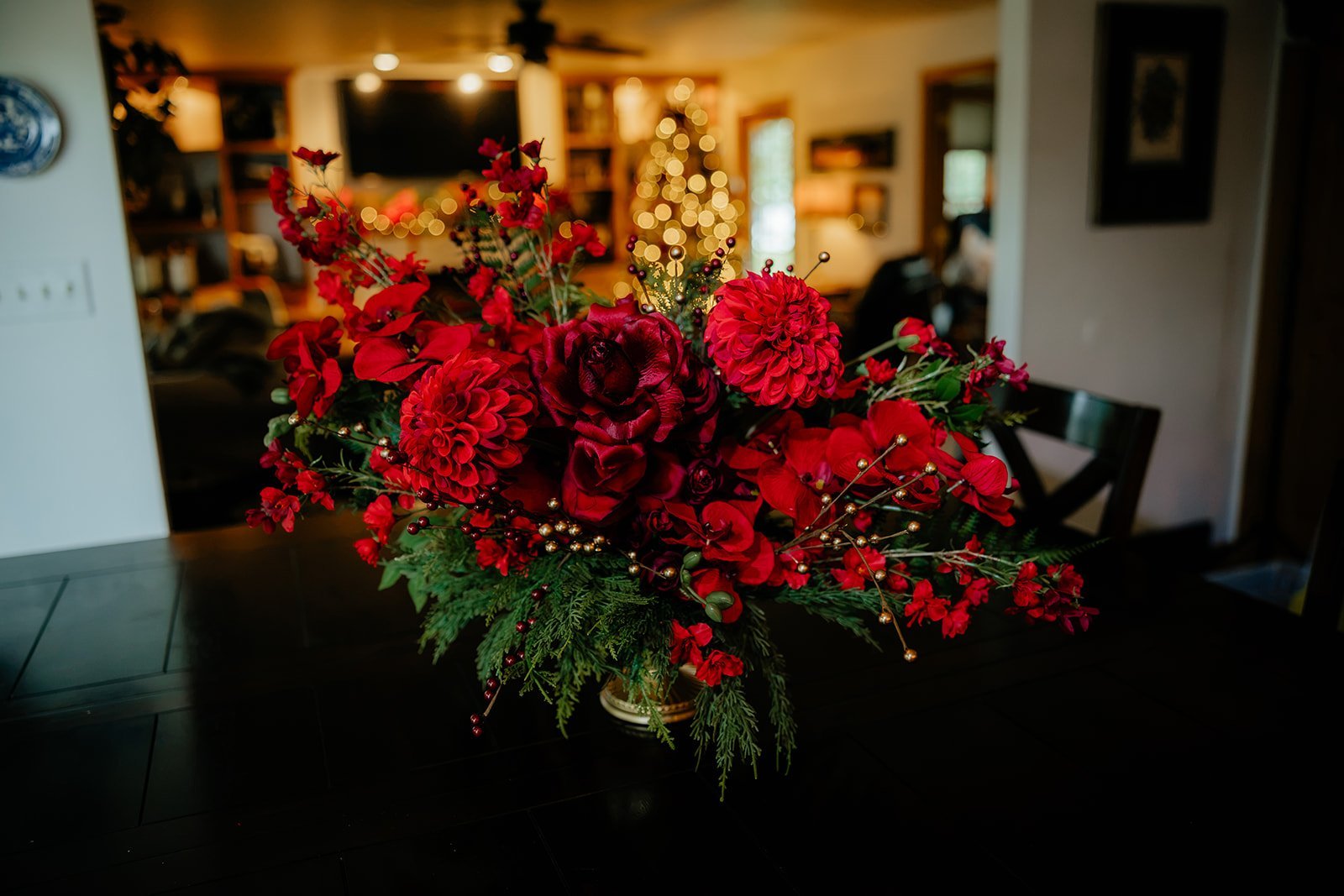 A vibrant red holiday floral arrangement with various red flowers, greenery, and decorative gold picks is placed on a dark table in a cozy, warmly lit living room decorated for Christmas, with a Christmas tree and a fireplace in the background.