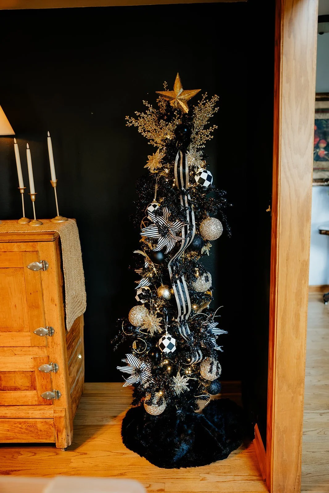 Decorated Christmas tree with black, silver, and gold ornaments, topped with a gold star, in a cozy living room corner.