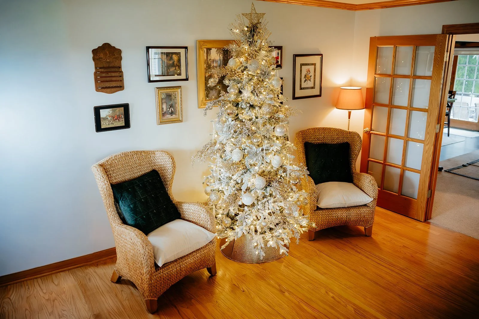 Decorated white Christmas tree in a living room with two wicker chairs, framed wall art, a floor lamp, and a wooden door leading to another room.