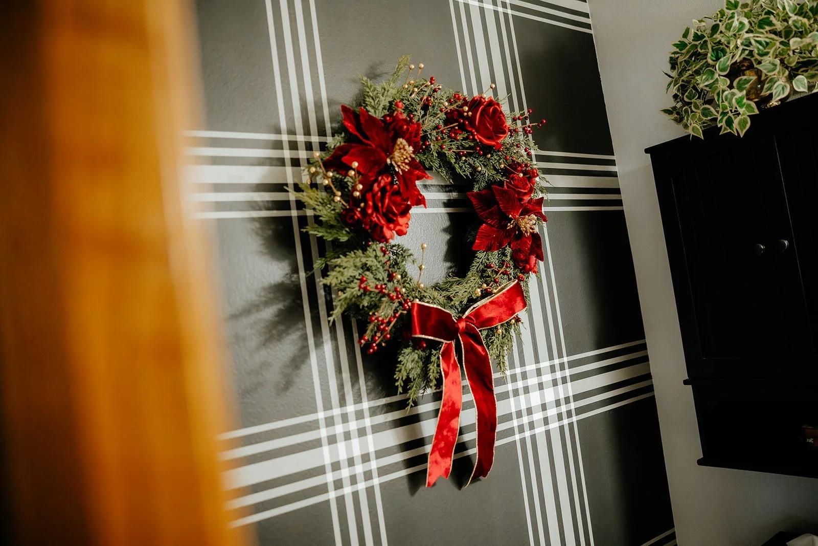 A Christmas wreath decorated with red poinsettias, red berries, and a red ribbon, hanging on a gray wall with white and black striped trim.