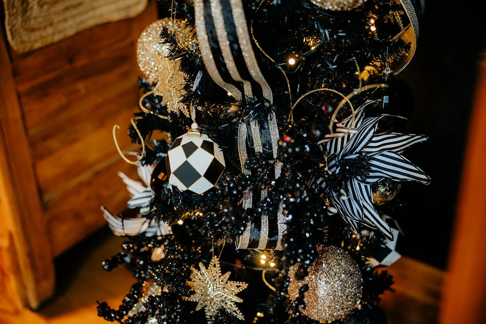 Close-up of a black Christmas tree decorated with black, gold, and white ornaments, including a striped butterfly, a geometric patterned ball, and a glittery gold ball, with warm lighting and wooden background.