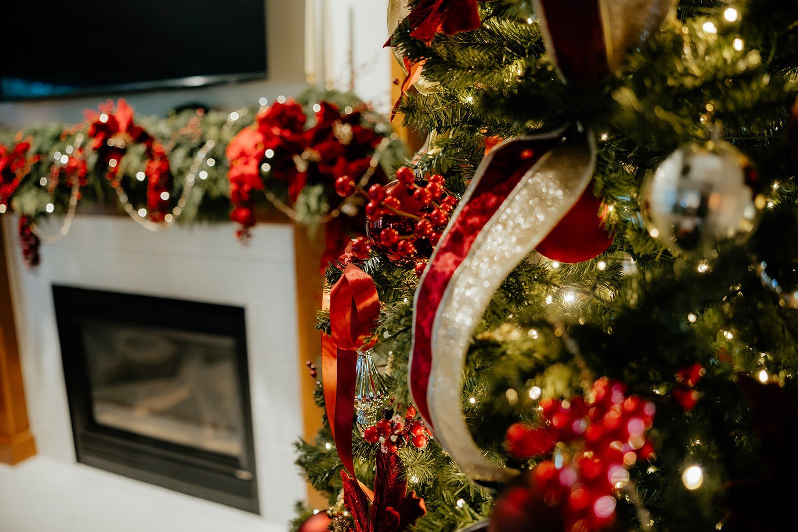 Close-up of a decorated Christmas tree with red and gold ornaments, ribbons, and string lights in a cozy room with a fireplace and holiday garland in the background.