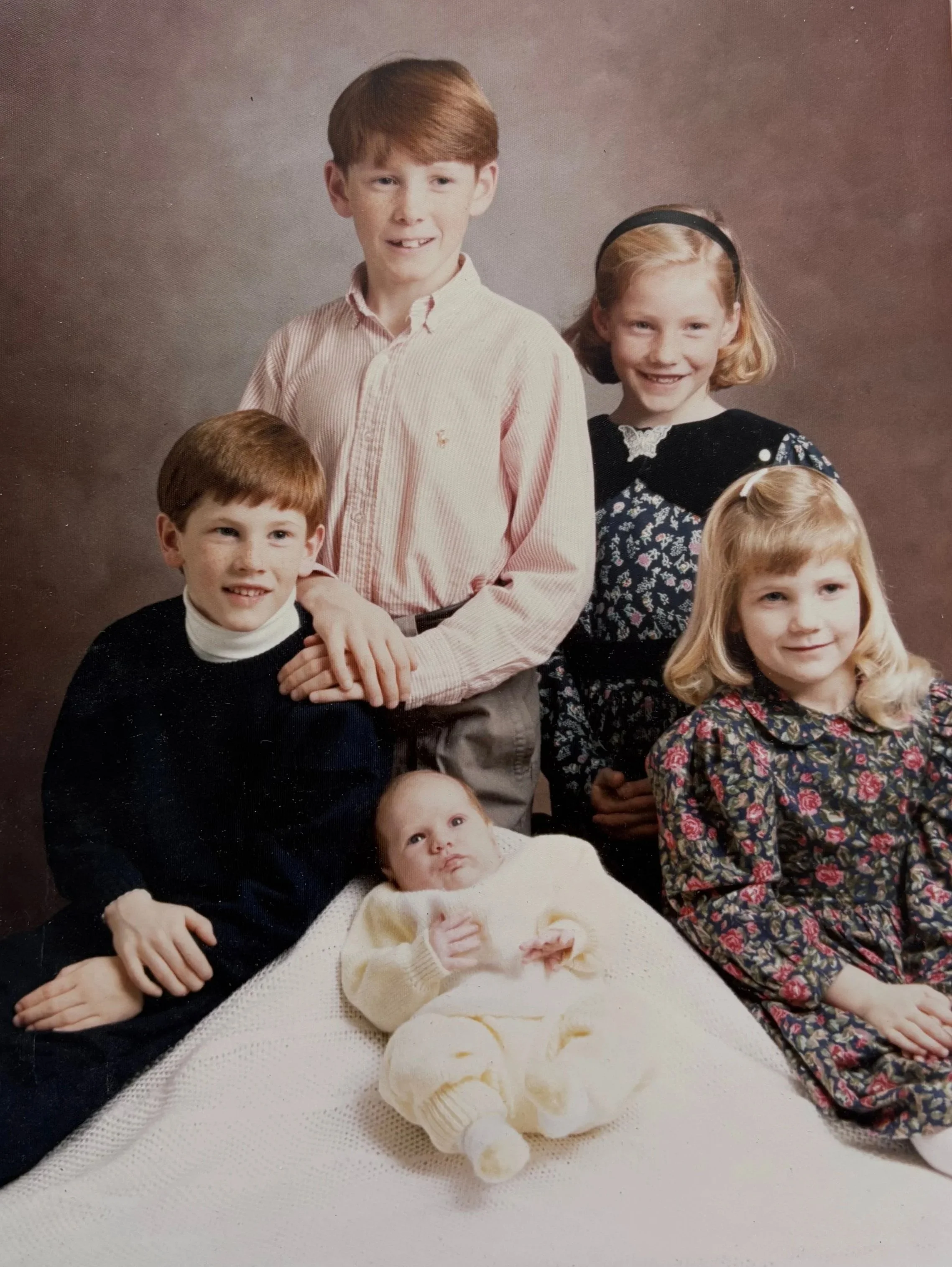 A family portrait featuring six children of varying ages, dressed in semi-formal clothing, posed against a neutral backdrop. The youngest child is lying on a white crocheted blanket in the foreground, while the older children are standing or sitting behind her.