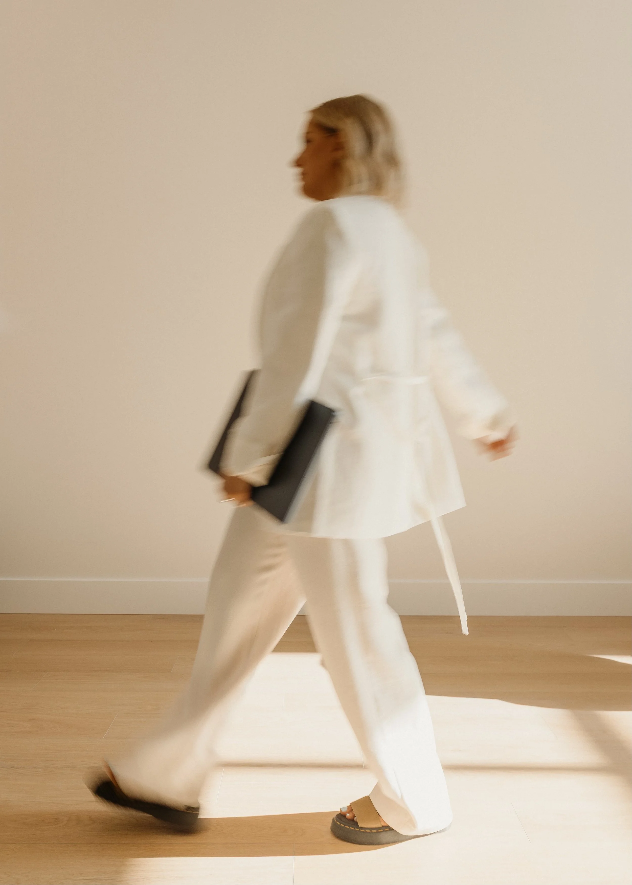 A person walking in a beige room, wearing a beige coat, wide-legged beige pants, and platform shoes. They are carrying a black book or folder and appear to be in motion.