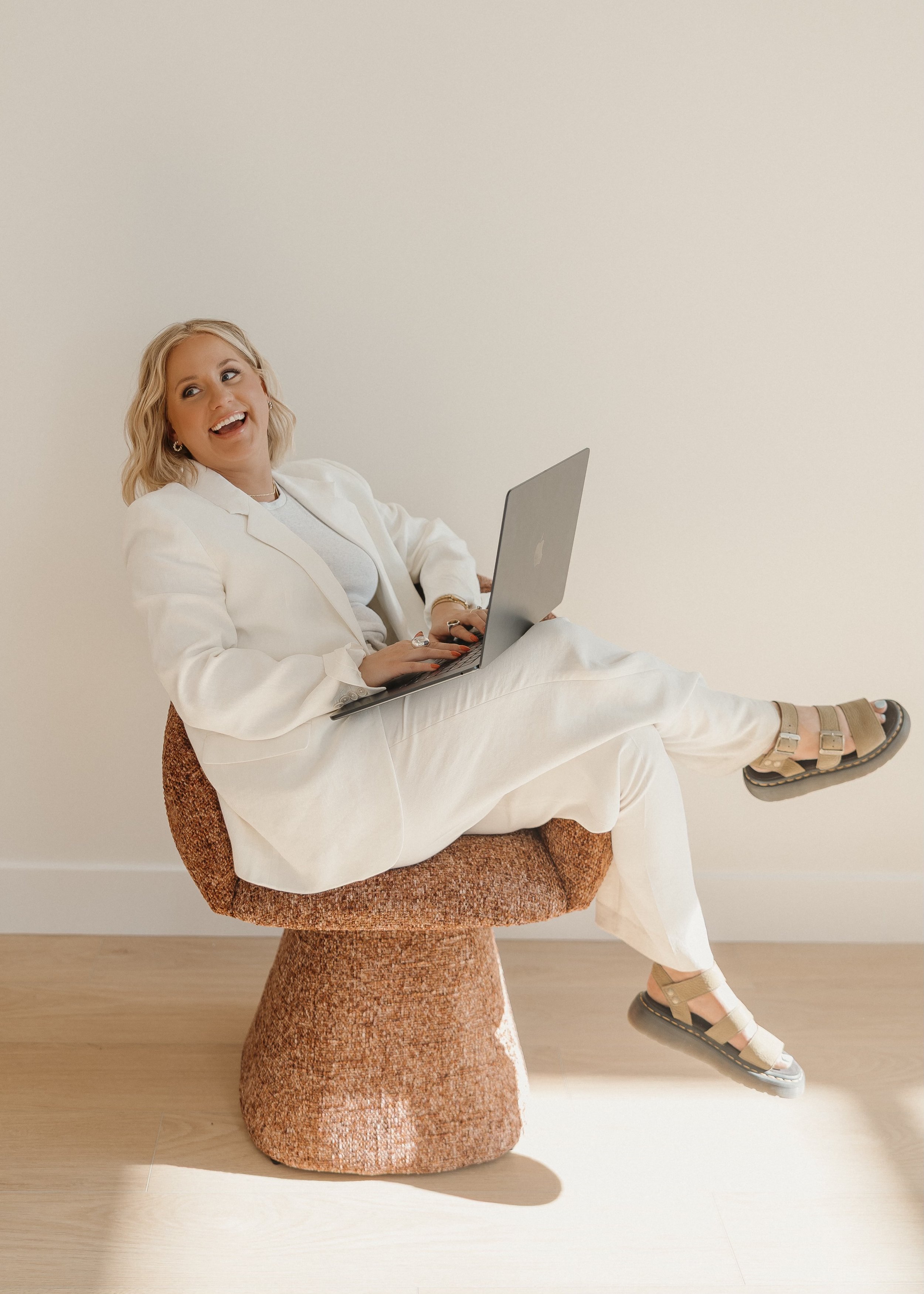 Woman with blonde hair sitting on a textured orange-brown chair, laughing while holding a silver laptop, dressed in a white suit and sandals with fringes, against a plain white wall.