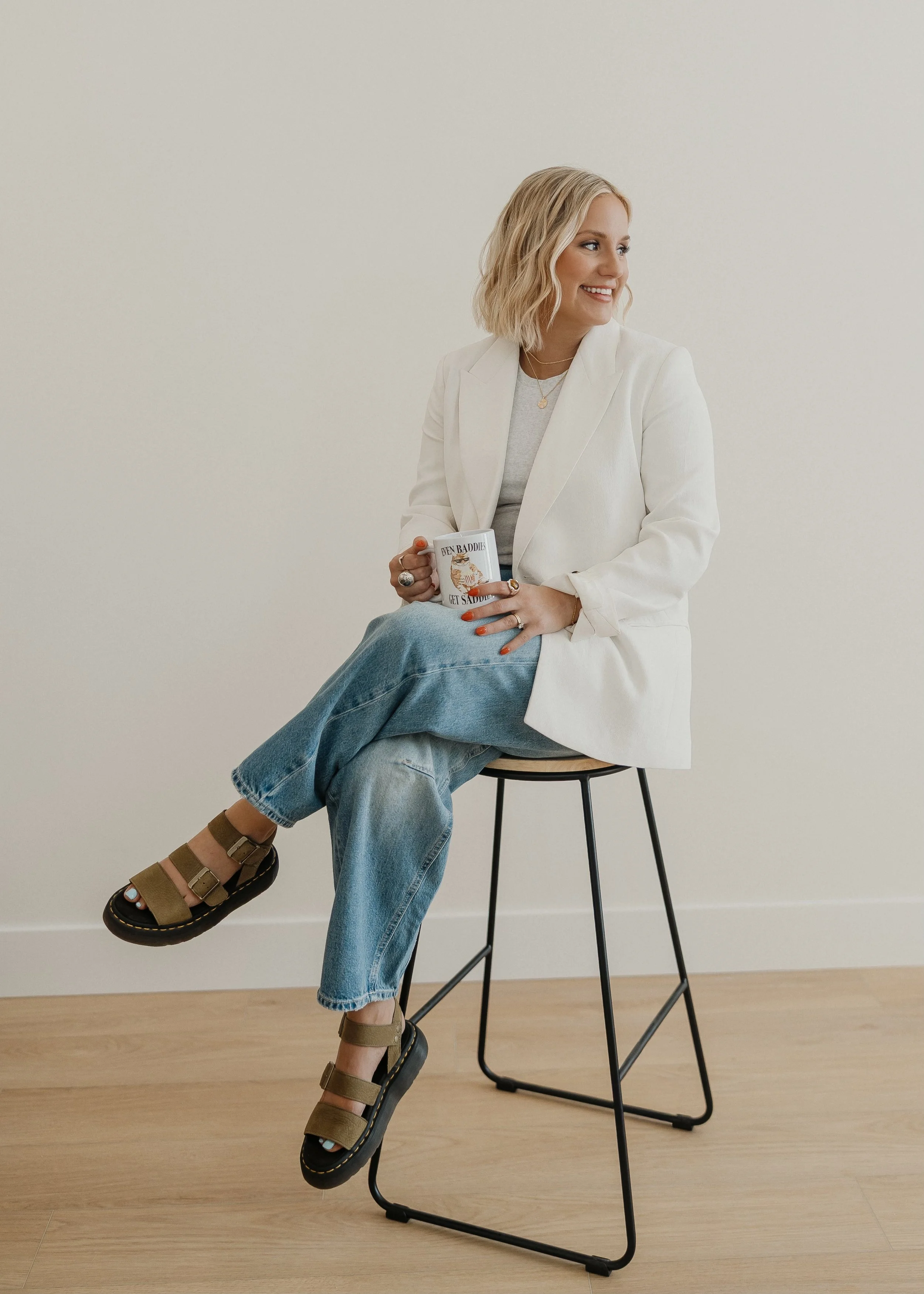 A woman with blonde hair sitting on a black stool against a plain light-colored wall. She is smiling and looking to her right, holding a white mug in her right hand and resting her left hand on her lap. She is wearing a white blazer, light gray top, baggy blue jeans, and brown sandals.