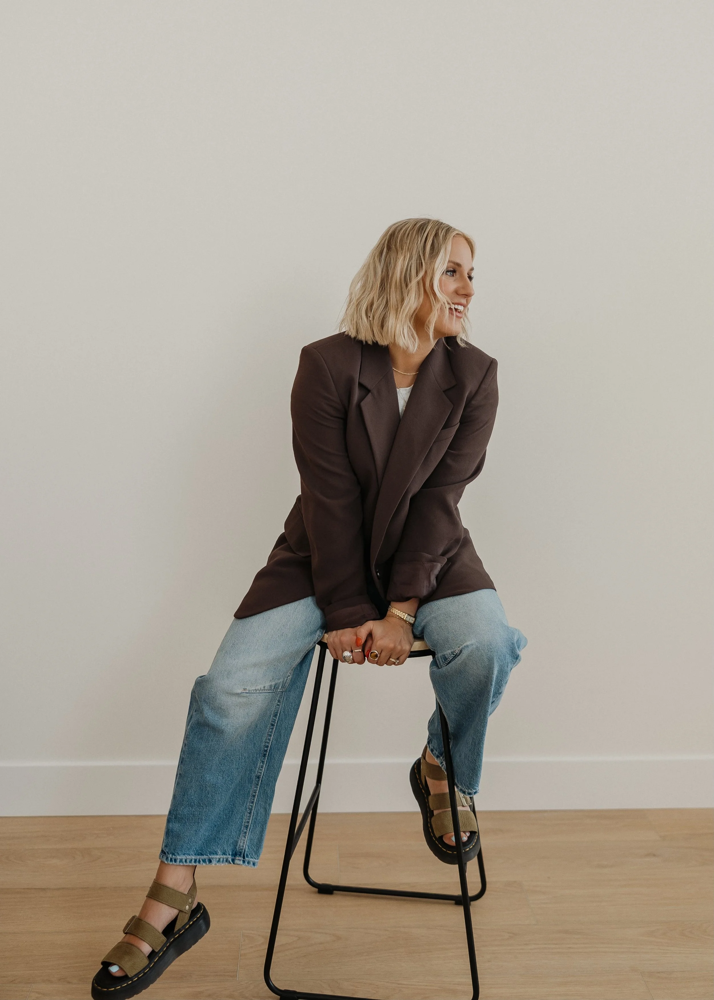 A woman with blonde wavy hair smiling and sitting on a black stool, wearing a dark brown blazer, light blue jeans, and brown platform sandals indoors against a plain white wall.