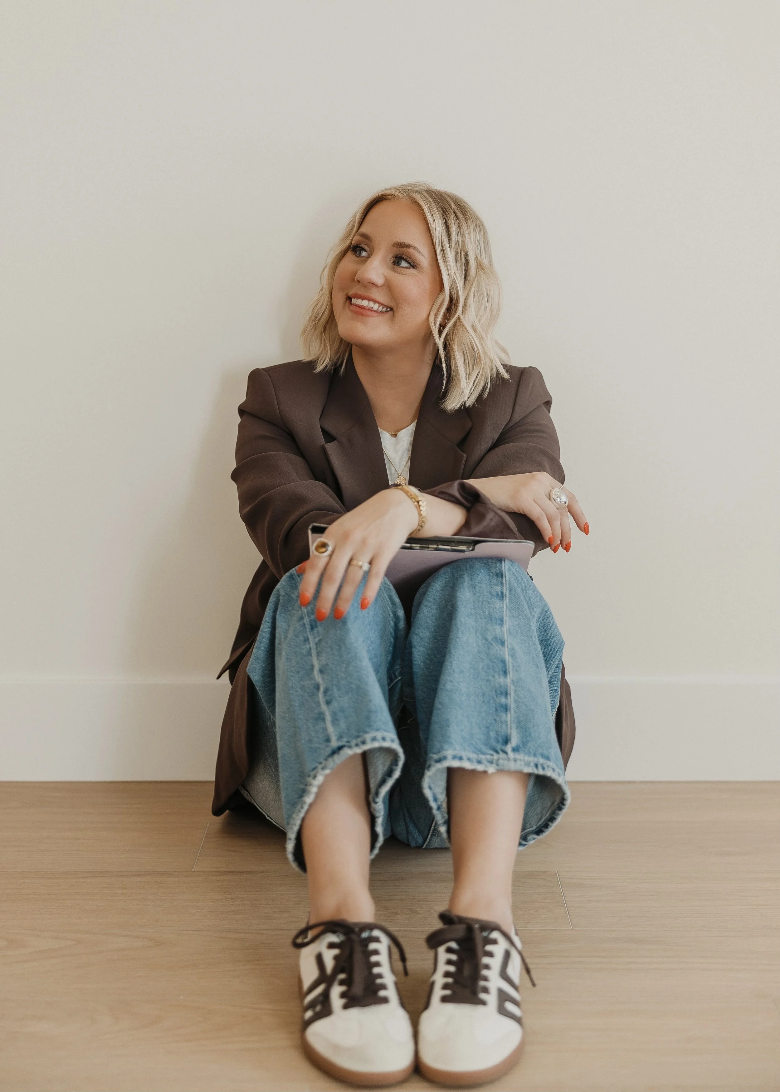A smiling woman sitting on the floor, wearing a brown blazer, blue jeans, and white sneakers with black accents, against a plain white wall.