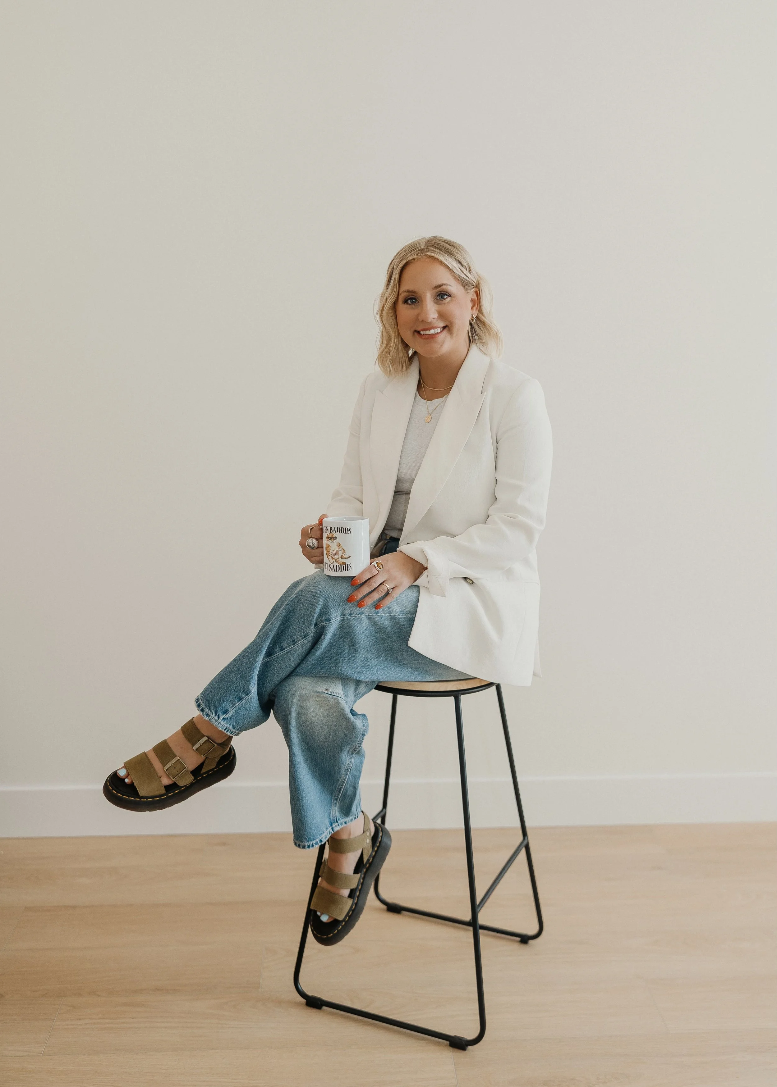 A woman sitting on a tall stool, holding a coffee mug, smiling, wearing a white blazer, beige sandal flats, and blue jeans, against a plain off-white wall.