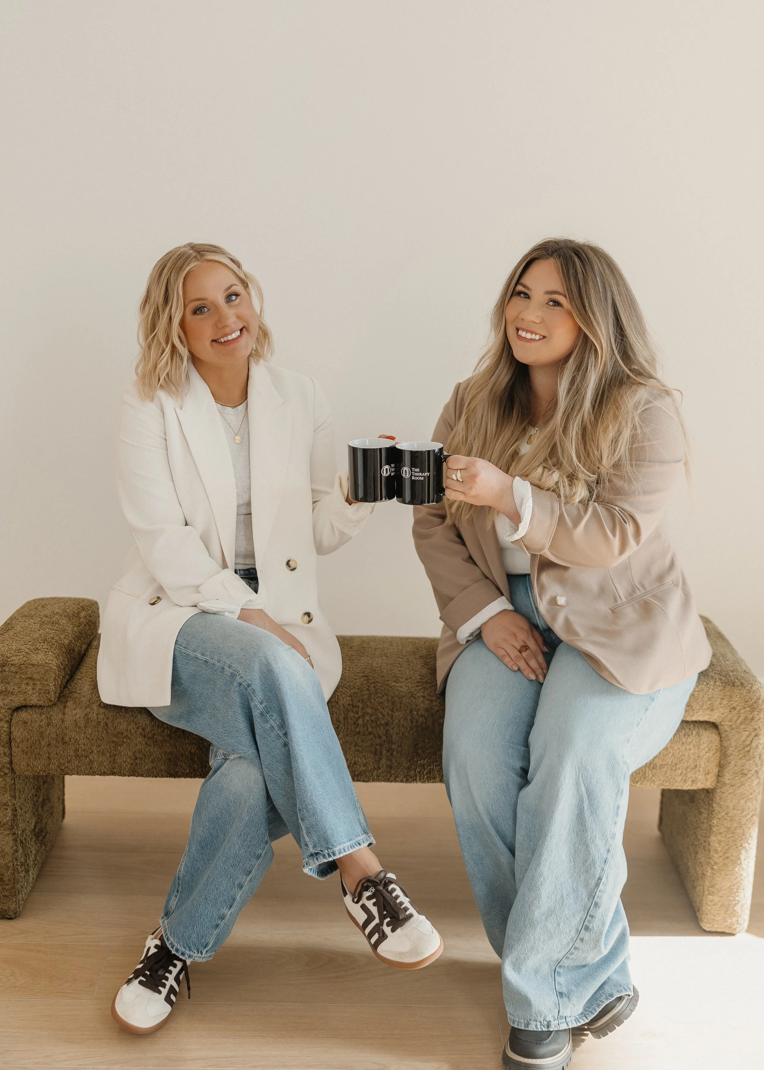 Two women sitting on a brown couch with a plain off-white wall behind them, smiling and holding black mugs that read "The Therapy Room." Both women are dressed casually in jeans and neutral-tone blazers, with the woman on the right wearing gray boots.