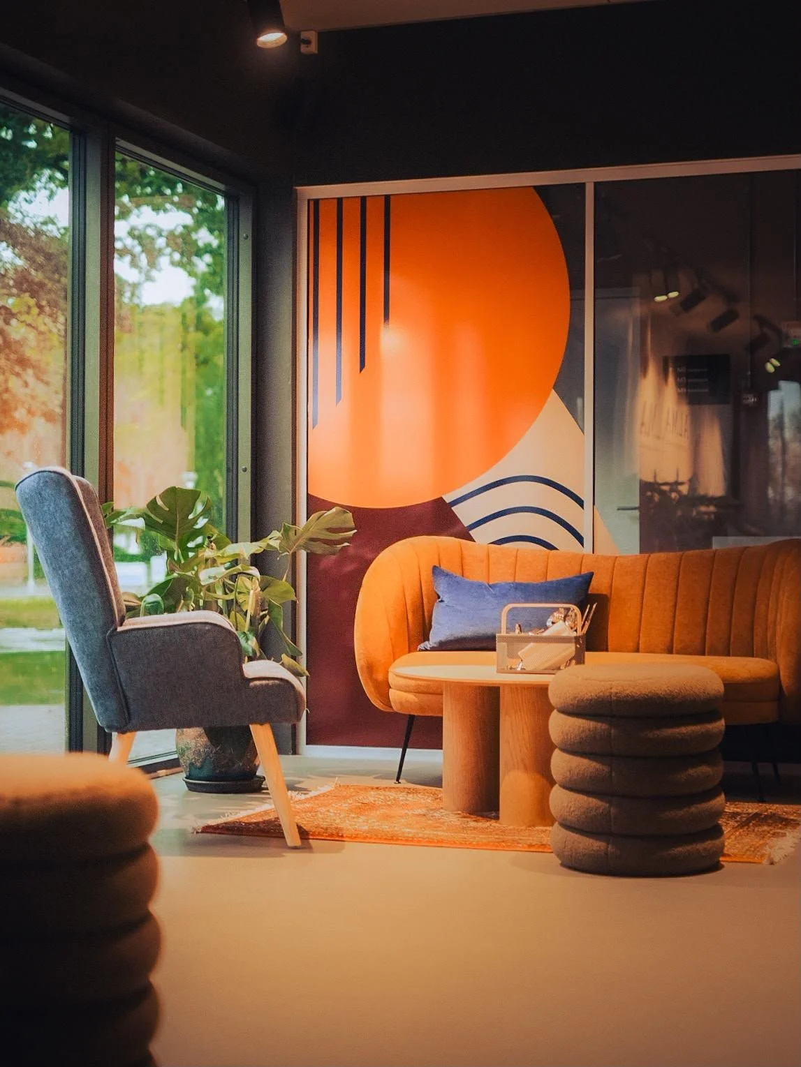 Interior of the lounge section of ALMA Cafe in Billund. Freen plants, a blue armchair, an orange sofa with a blue pillow, a wooden coffee table with a tissue box, and abstract wall art with orange, black, beige, and blue shapes.